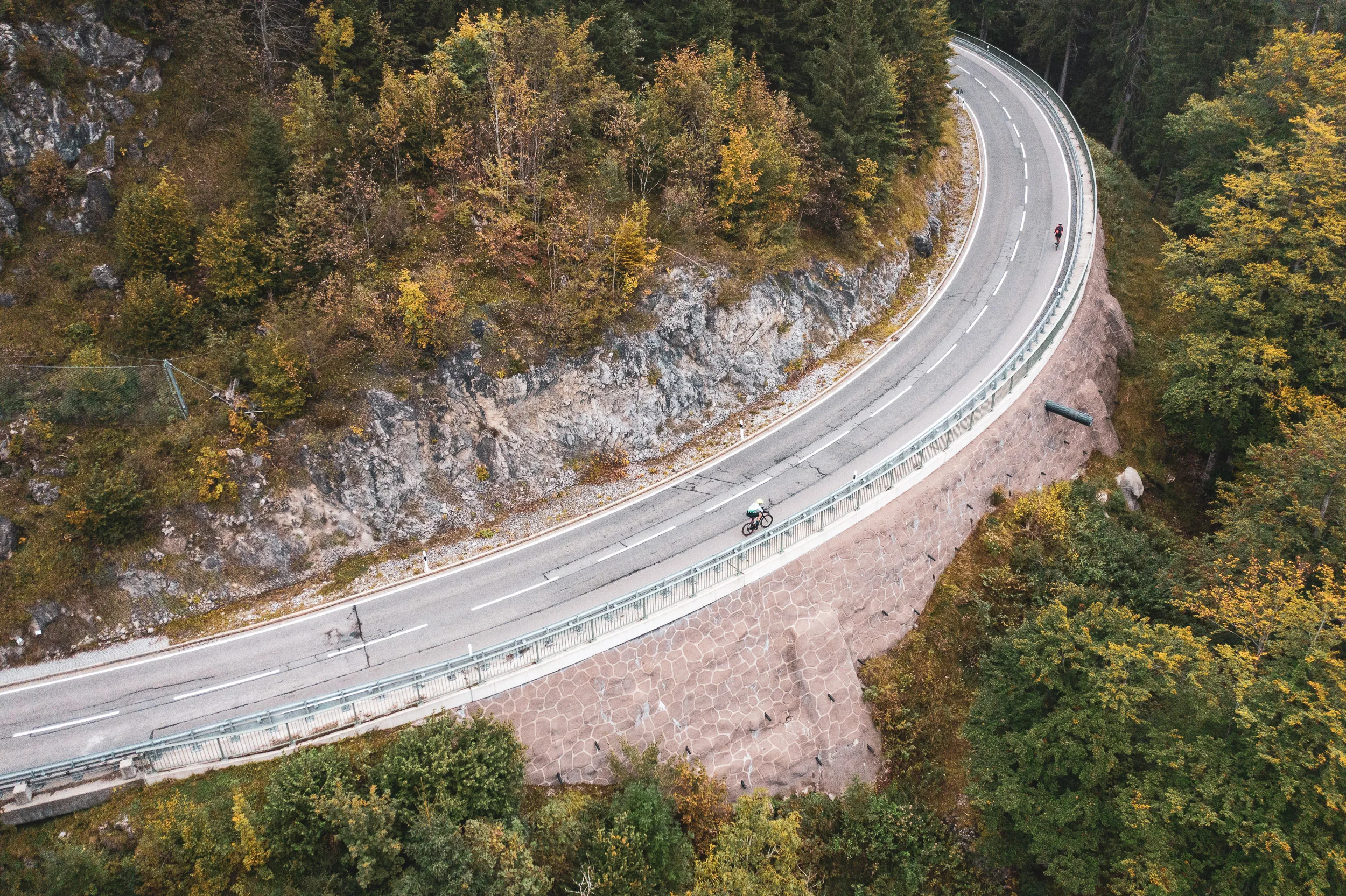 Zwei Rennradfahrer folgen einer engen Kurve zwischen Felsen und herbstlichem Wald am Oberjoch.