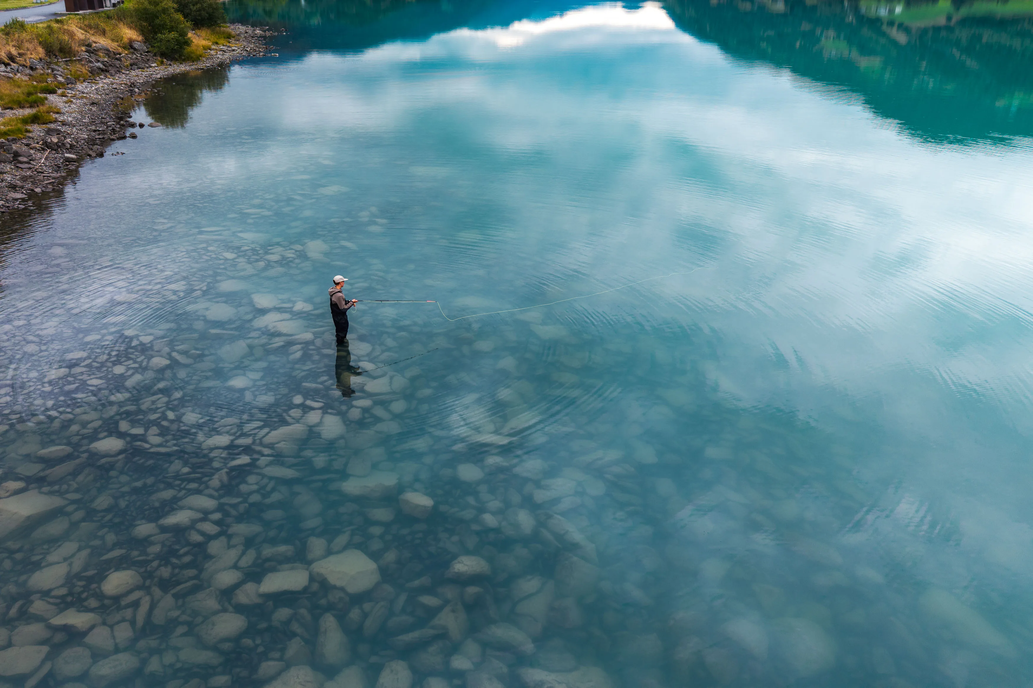 Angler steht in einem klaren Bergfluss, dessen Oberfläche Wolken und Berghänge spiegelt
