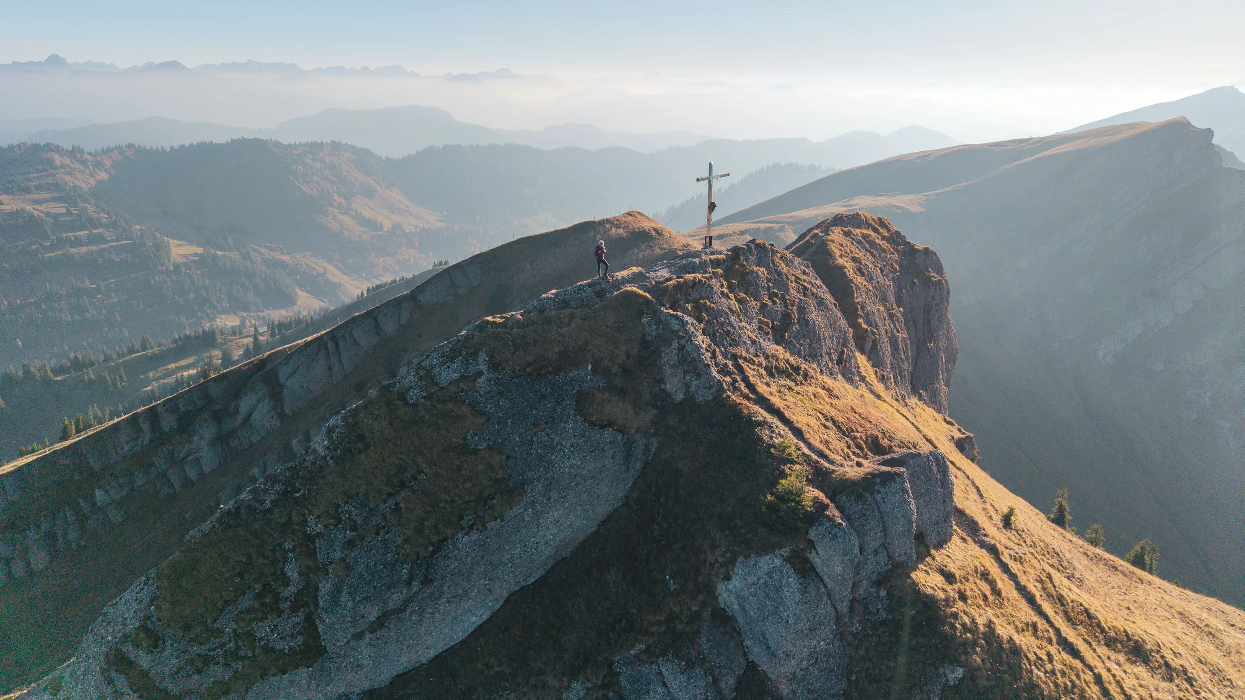 Eine Wanderin steht am Gipfelkreuz auf einem schmalen Grat im späten Licht