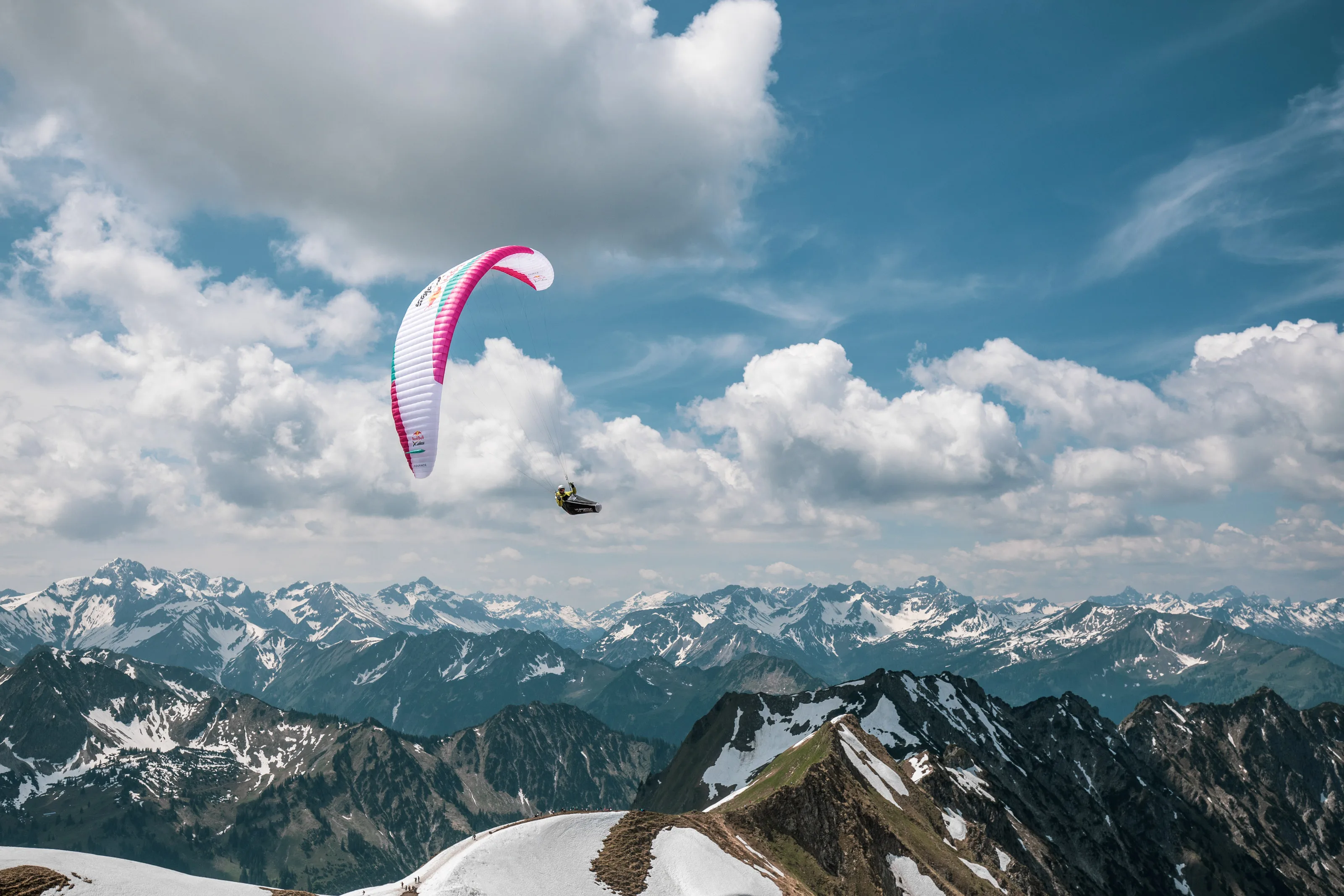 Michael Lacher fliegt unter einem pinkweißen Gleitschirm vor Wolken und verschneiten Gipfeln.