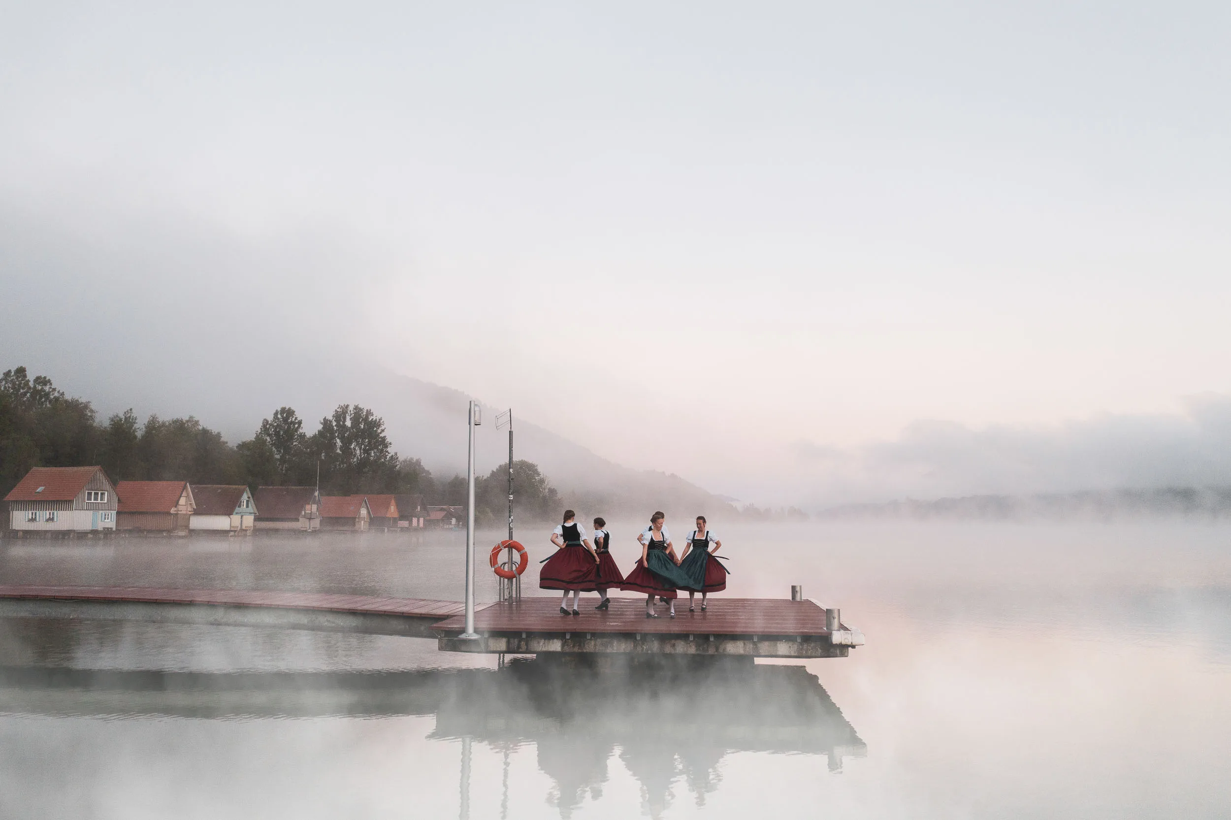 Tänzerinnen in Tracht auf einem Steg am stillen Alpsee im Morgennebel.