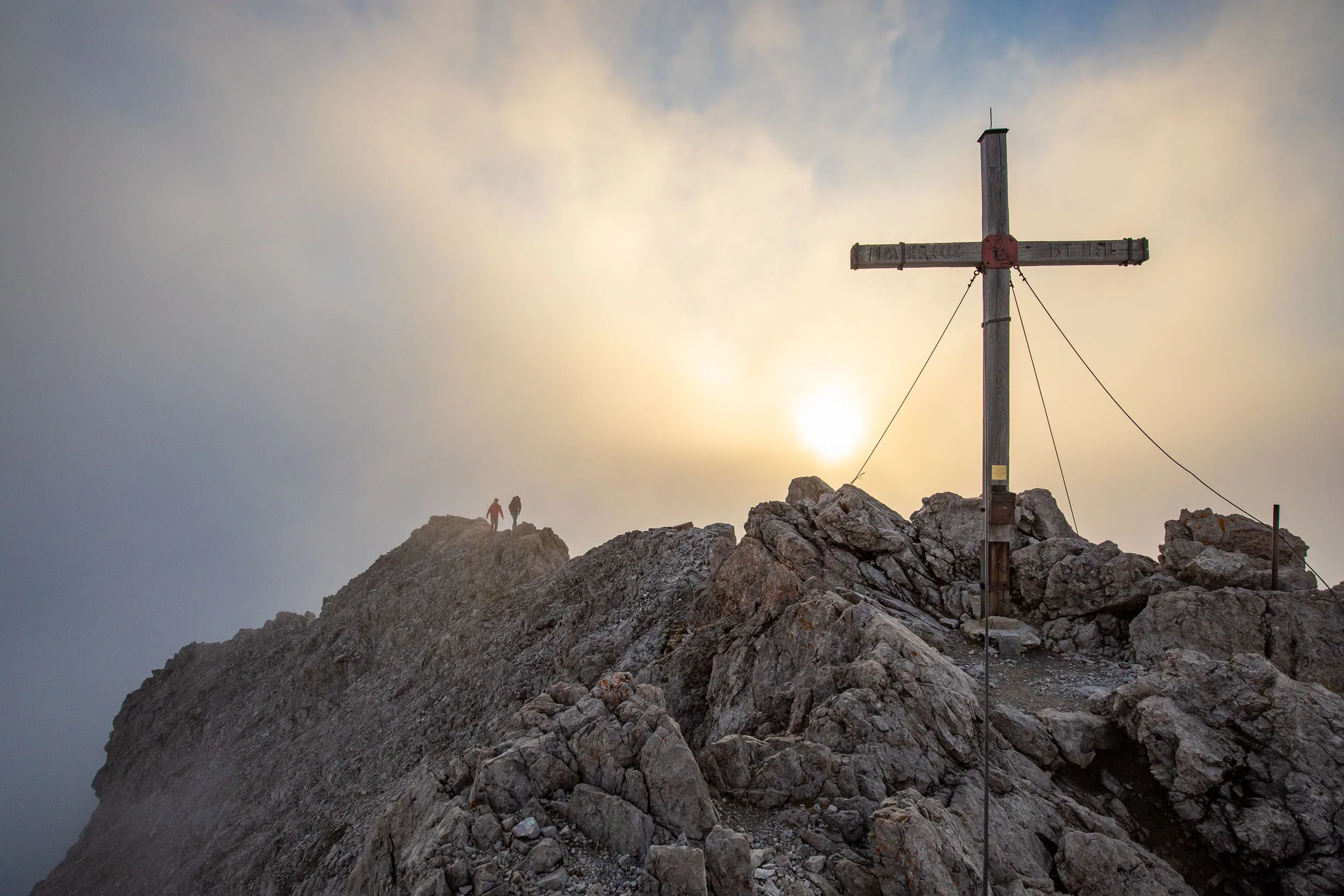 Ein Gipfelkreuz steht auf hellem Kalkgestein vor der tiefen Sonne