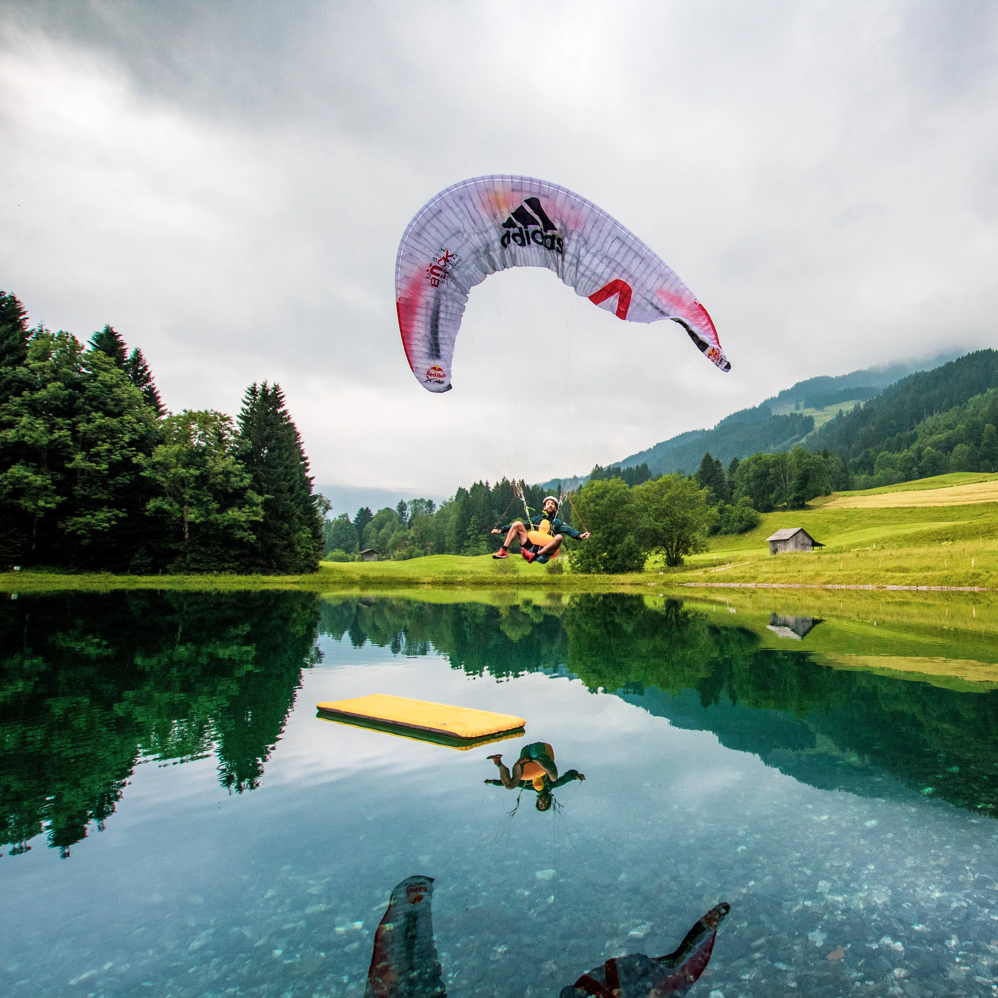 Manuel Nübel schwebt mit Gleitschirm und Badeente über einem Teich; sein Spiegelbild liegt im Wasser.