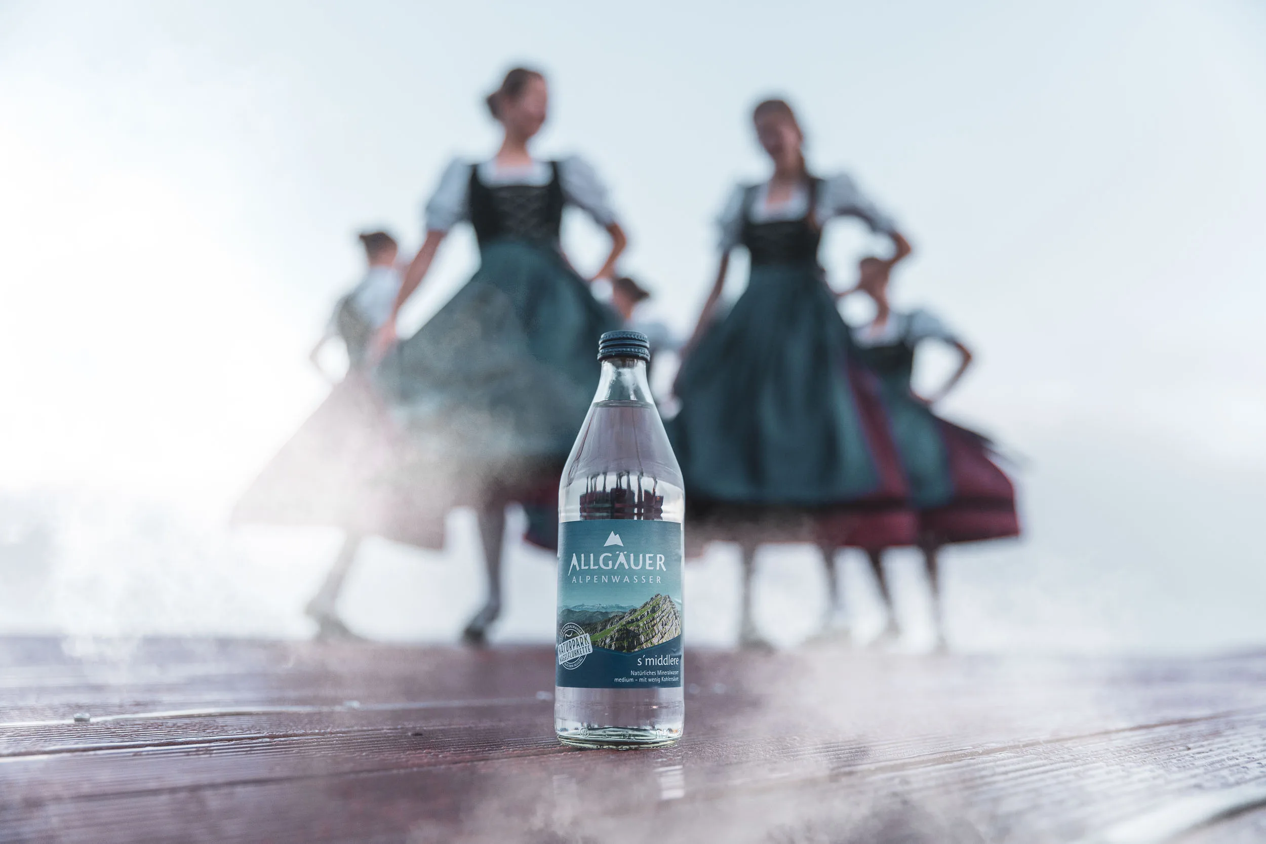 Wasserflasche im Vordergrund, dahinter Tänzerinnen in Tracht auf einem Steg am Alpsee.