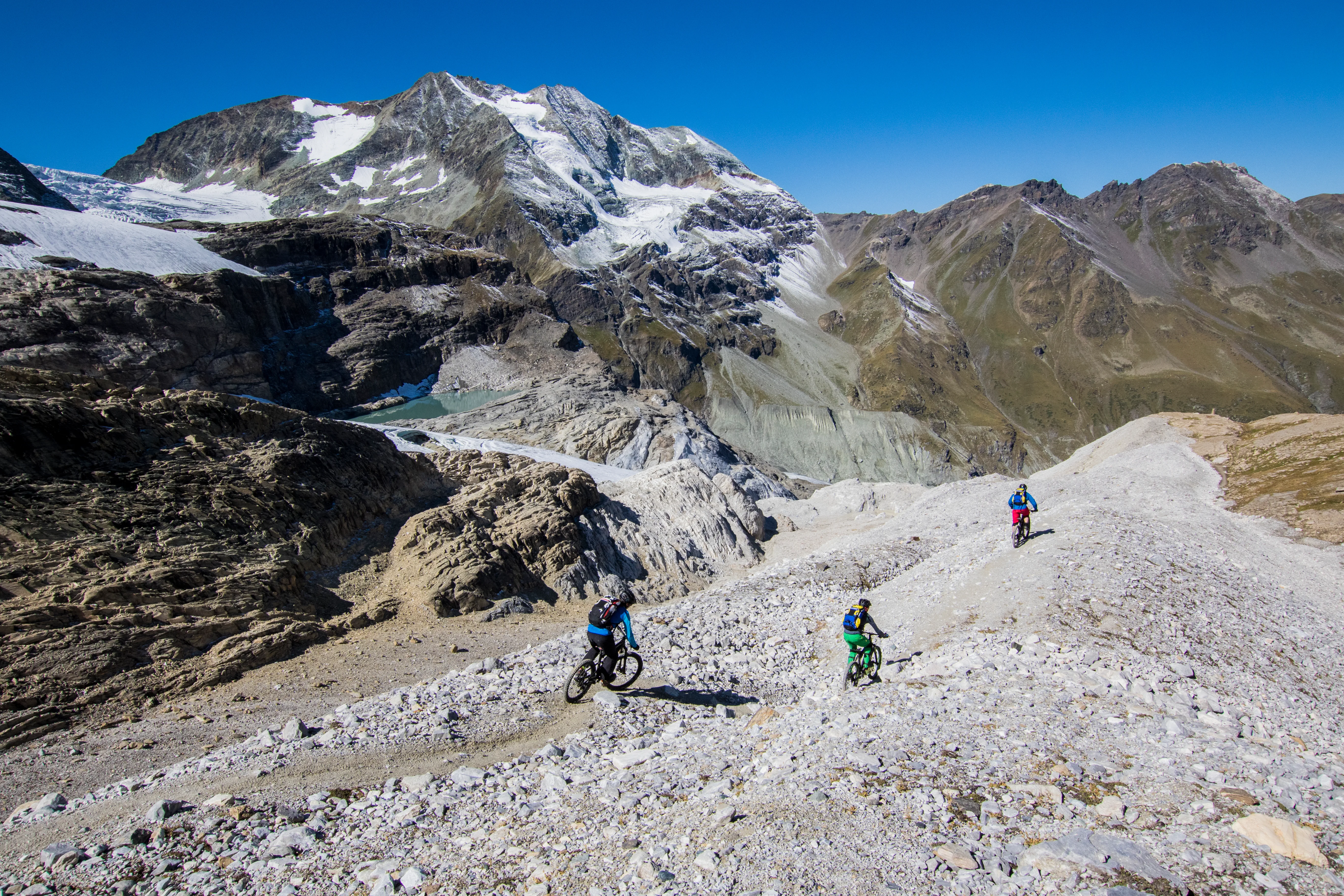 Robert Härle, Florian Bergmann und Florian Häusler fahren auf einem schmalen Trail vor vergletscherten Gipfeln.