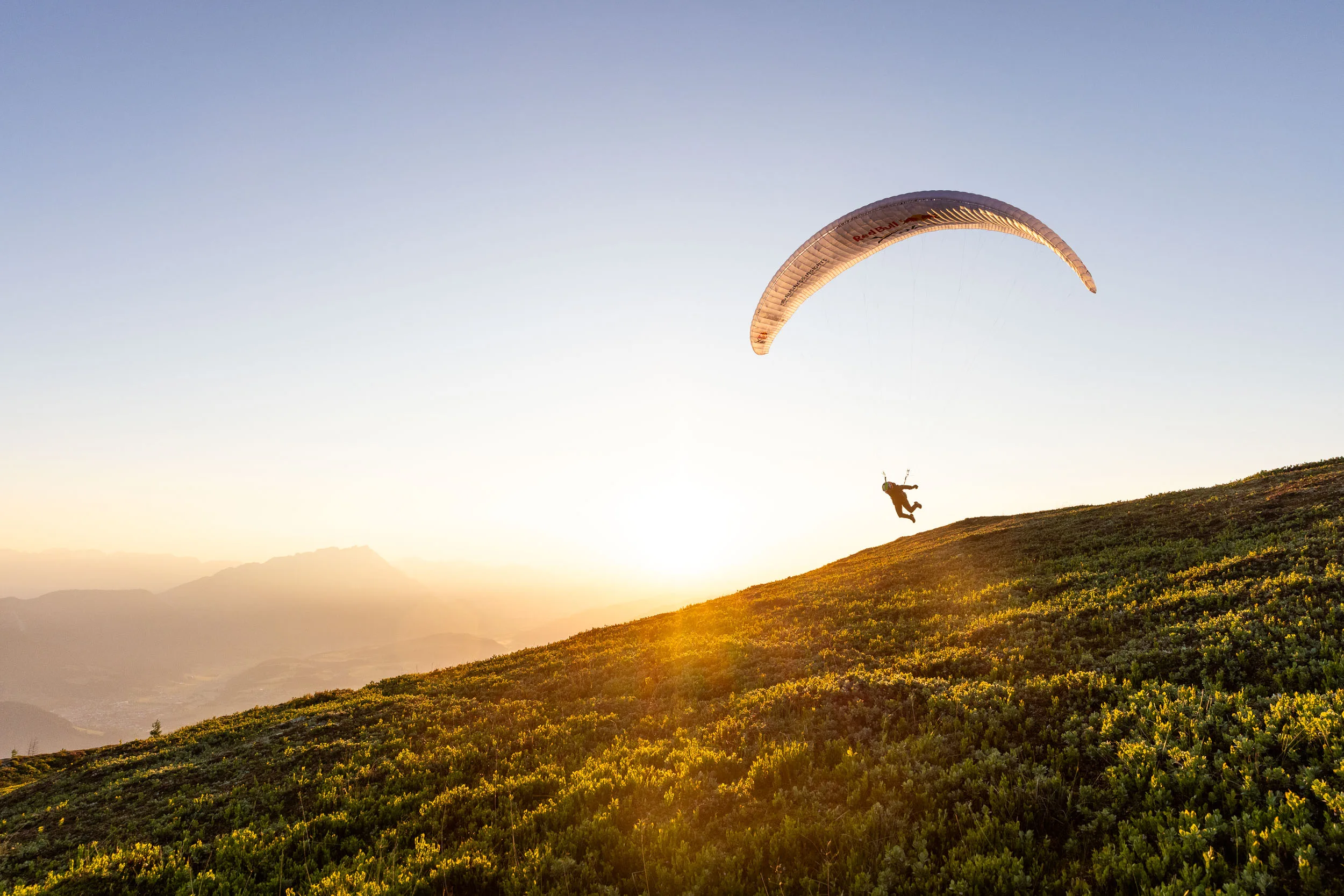Gleitschirmpilotin mit Gleitschirm ueber einem Hang im Ennstal bei Sonnenaufgang
