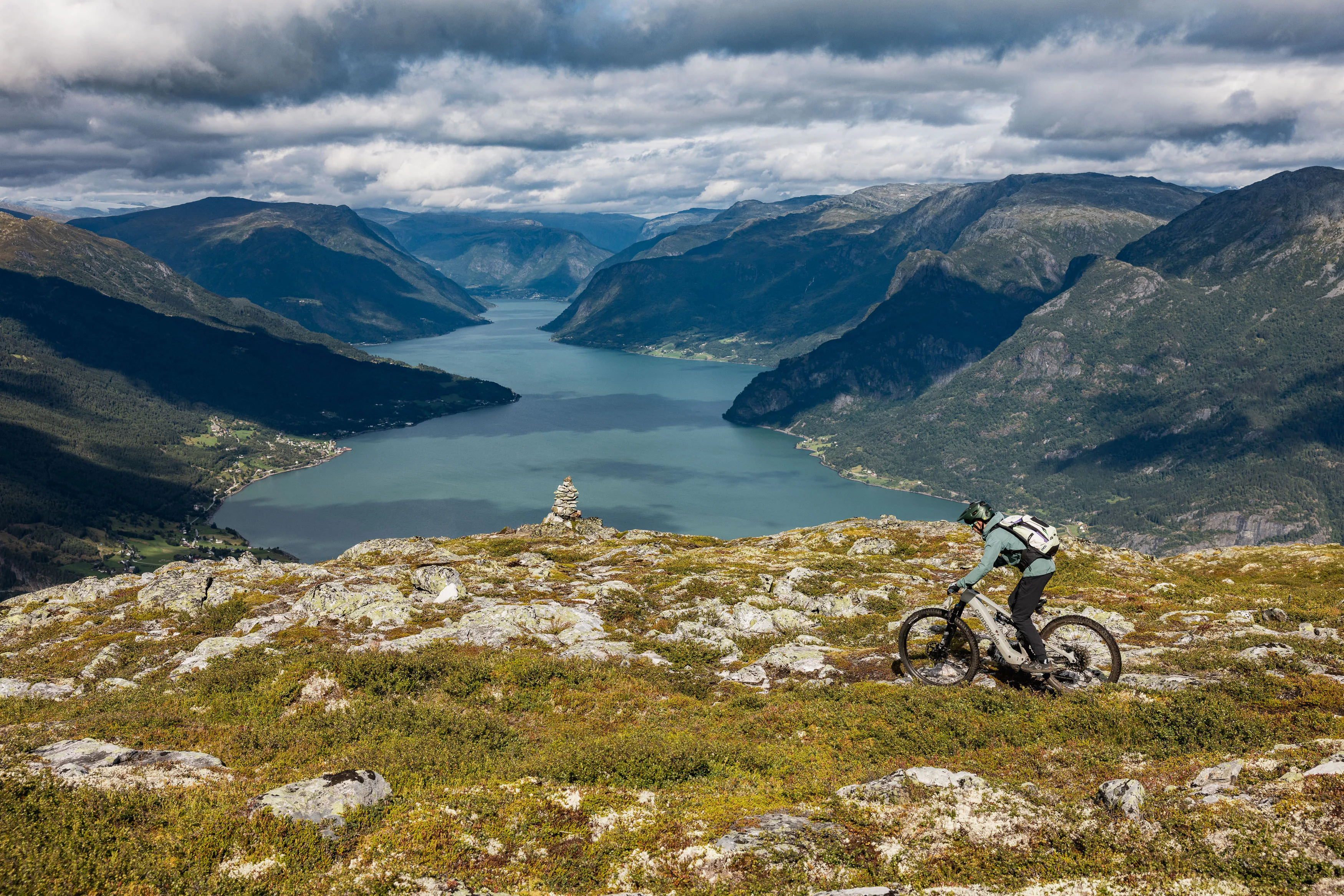 Mountainbiker fährt über felsiges Gelände vor einem tiefen Fjordtal unter dunklen Wolken