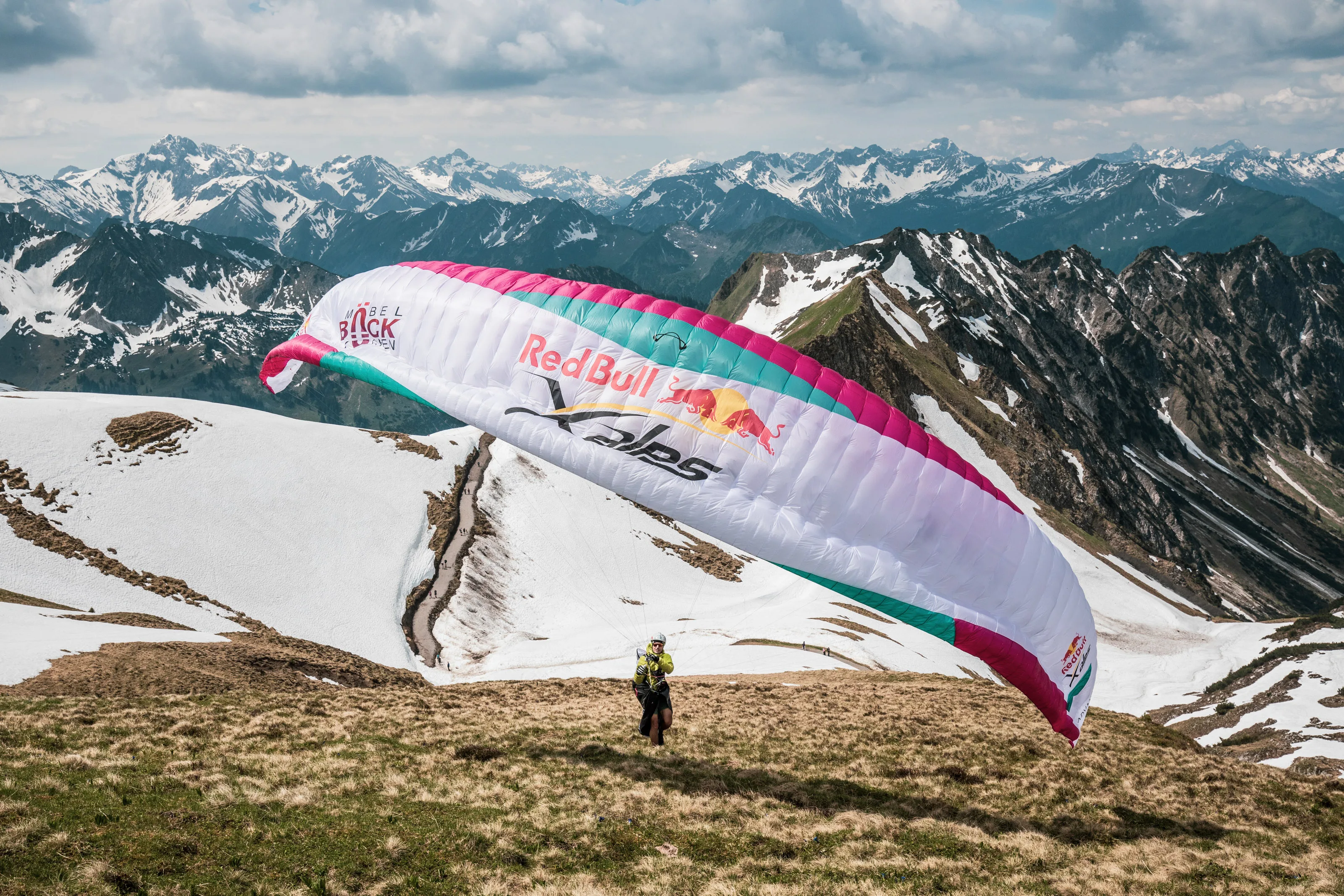 Ein pinkweißer Gleitschirm spannt sich knapp über einem schneefleckigen Hang am Nebelhorn.
