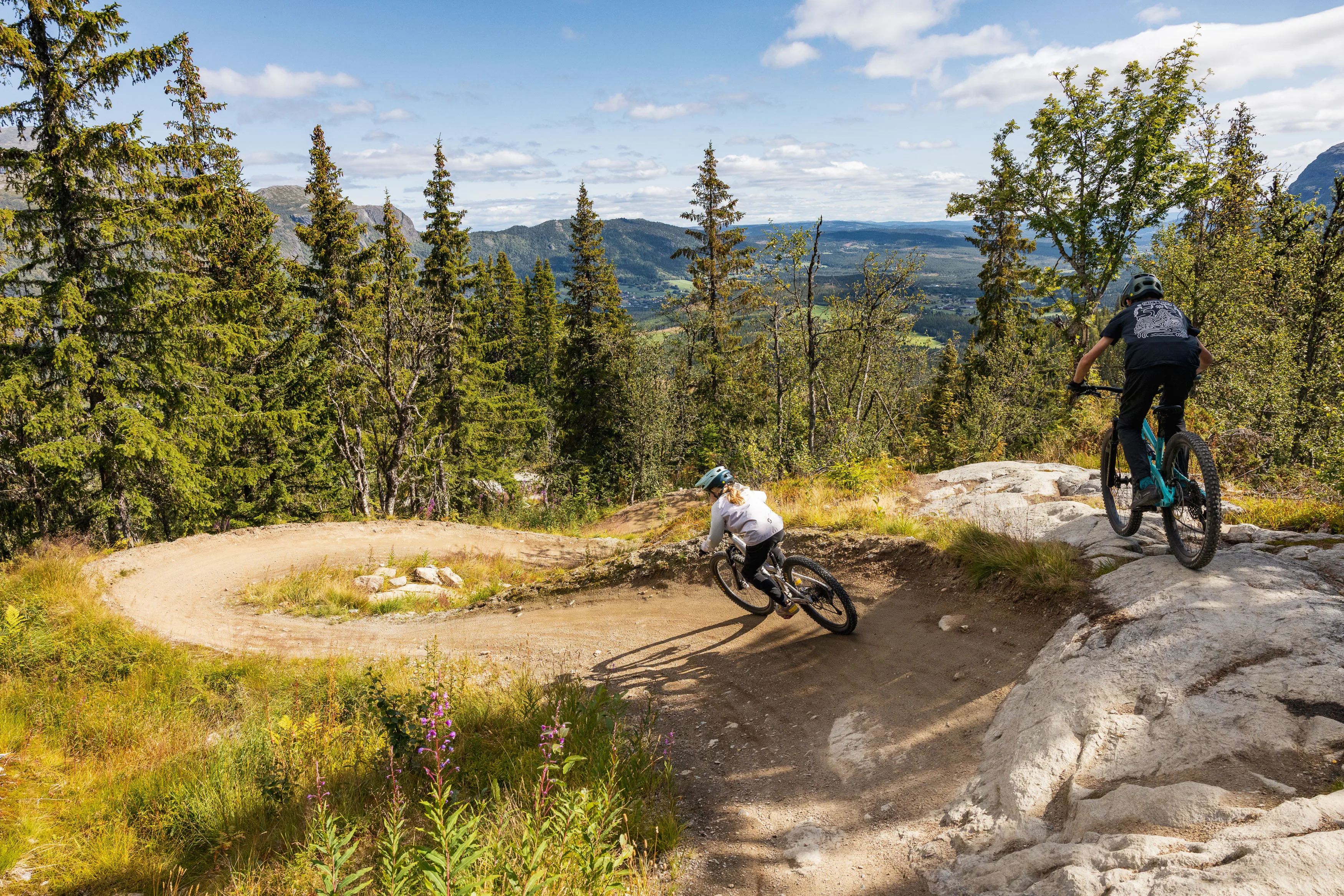 Zwei Mountainbiker nehmen versetzt eine steile Kurve zwischen Fichten und Felsen