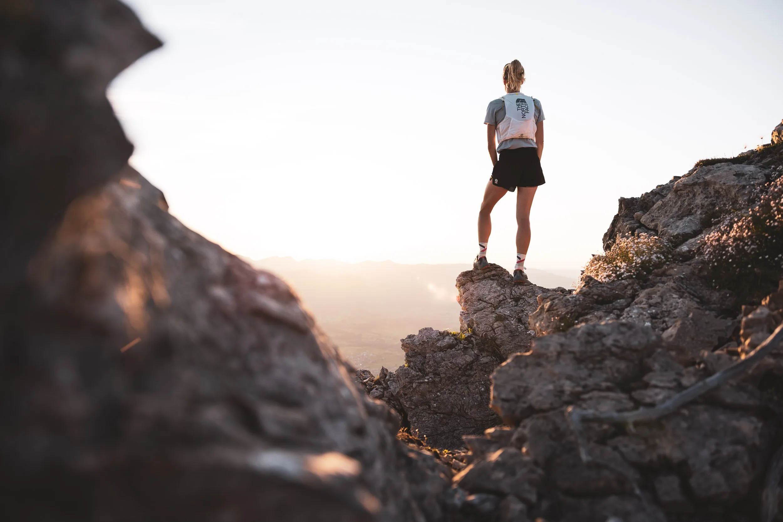 Trailrunnerin steht zwischen Felsen im hellen Gegenlicht über dem Tal