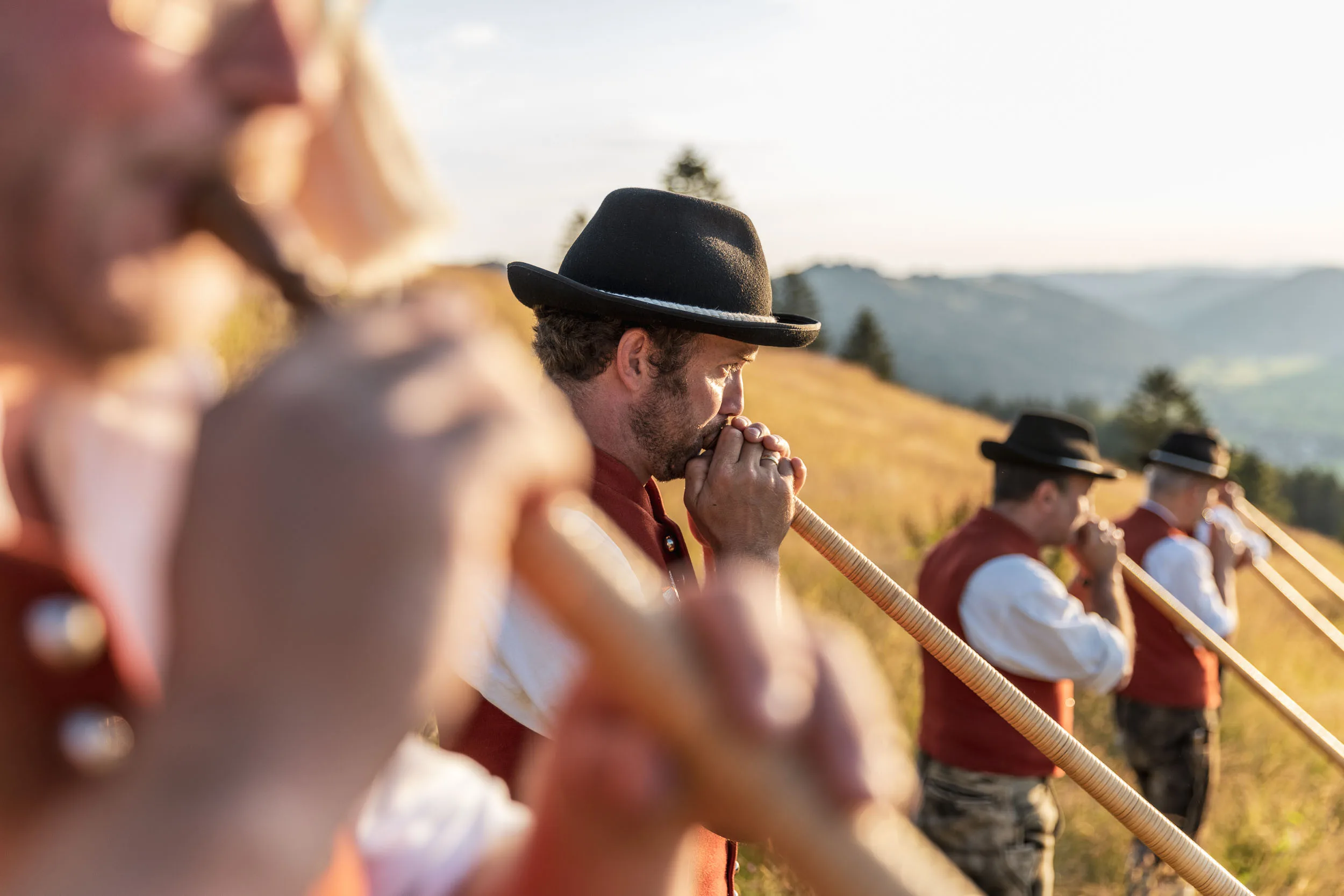 Alphornbläser in Tracht auf einer Bergwiese im Allgäu bei tief stehender Sonne.