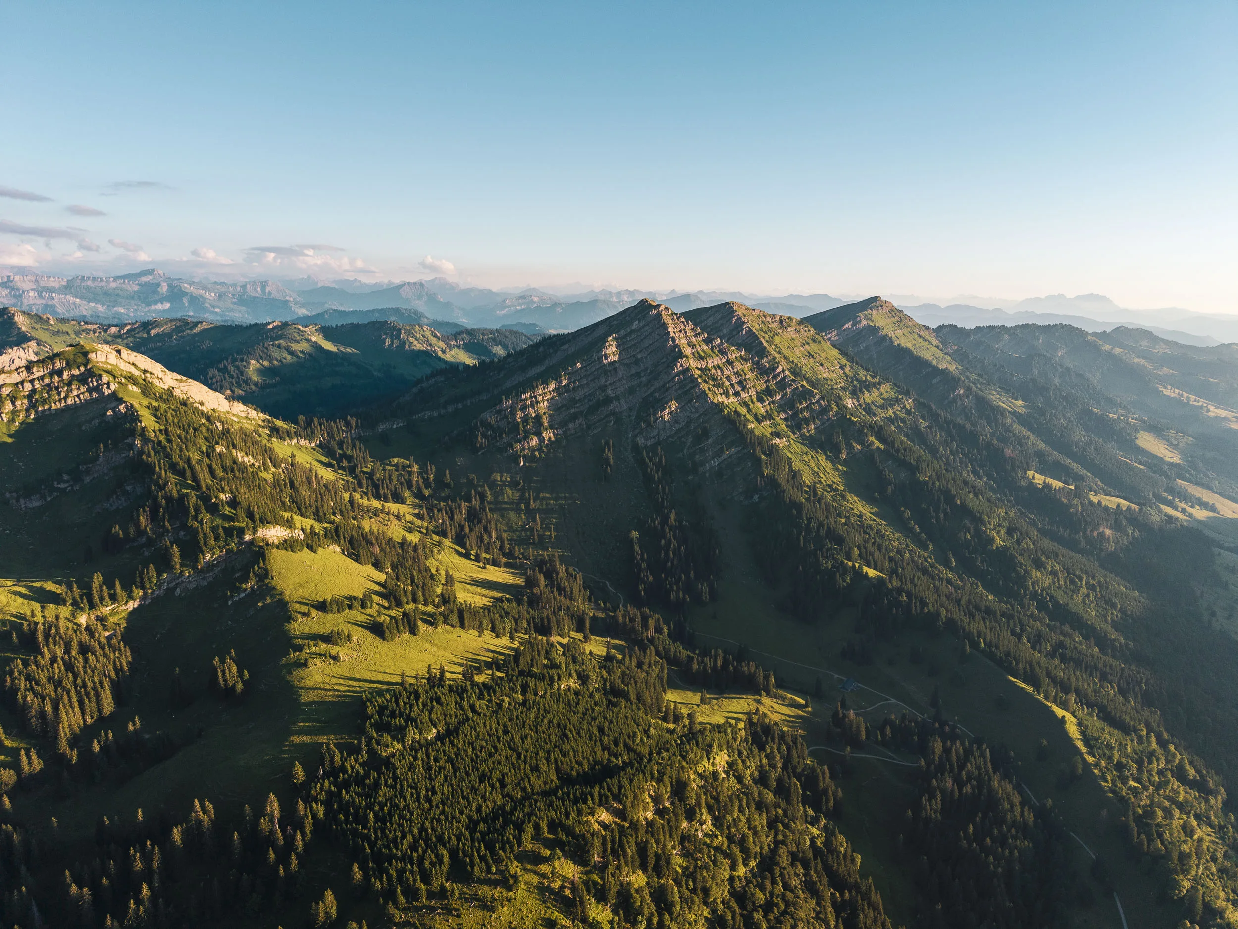 Sonnenlicht trifft grüne Bergflanken und Nadelwälder, während die Täler bereits im Schatten liegen.