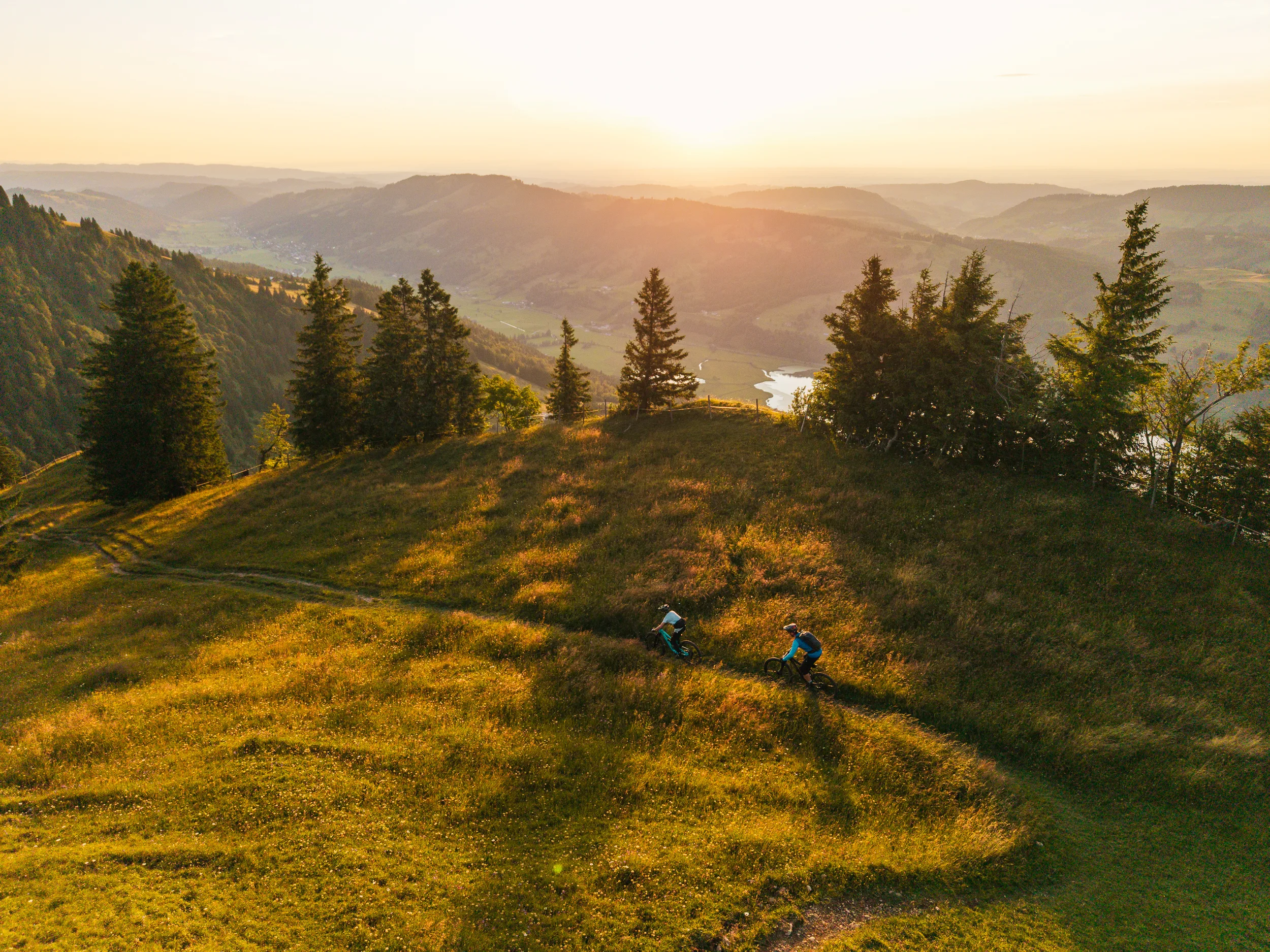Zwei Mountainbiker fahren über eine Bergwiese im warmen Licht der tiefen Sonne