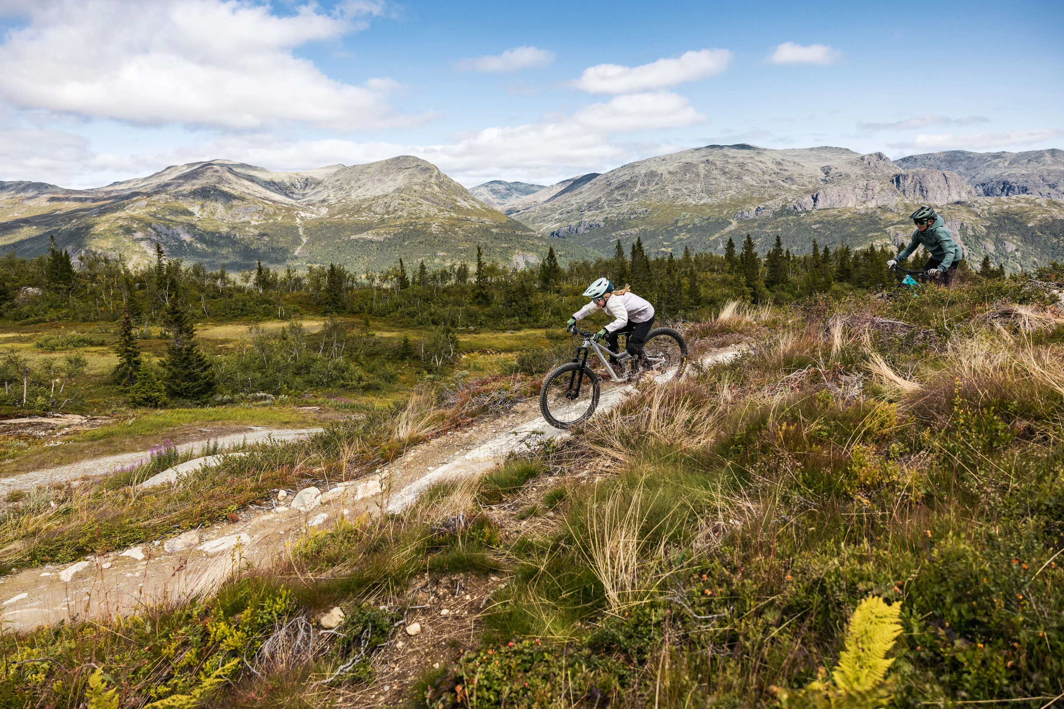 Zwei Mountainbiker folgen einander auf einem offenen Trail vor einer Bergkette