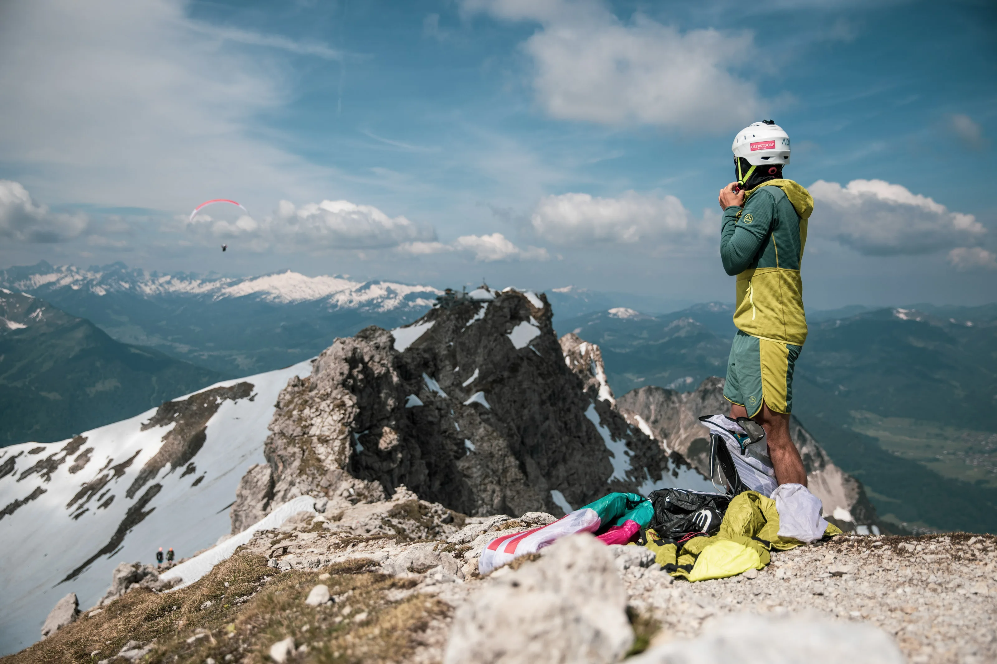 Mit Helm und Gleitschirmausrüstung steht Michael Lacher am Grat und blickt über die Berglandschaft.