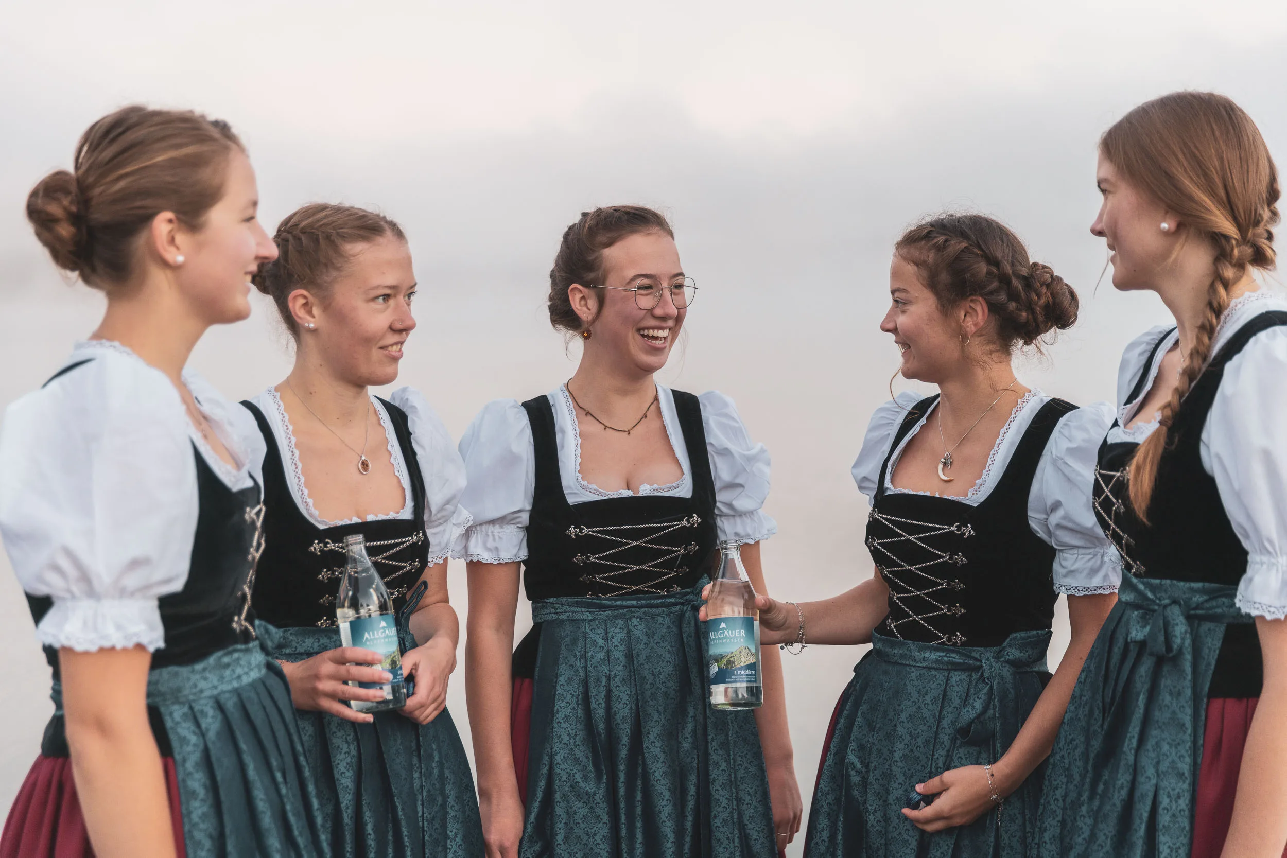 Tänzerinnen in Tracht unterhalten sich am Alpsee in weichem Morgenlicht.