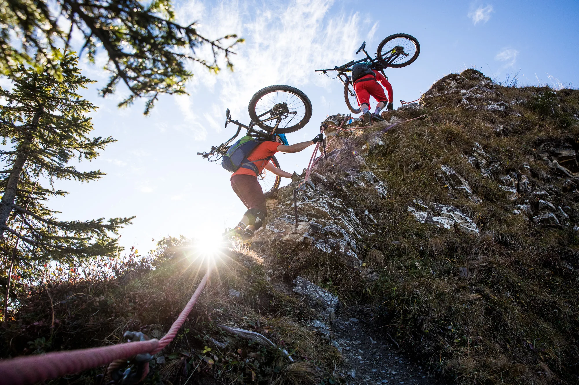 Mountainbiker tragen ihre Räder über einen Felsabsatz, während die Sonne durch die Bäume scheint