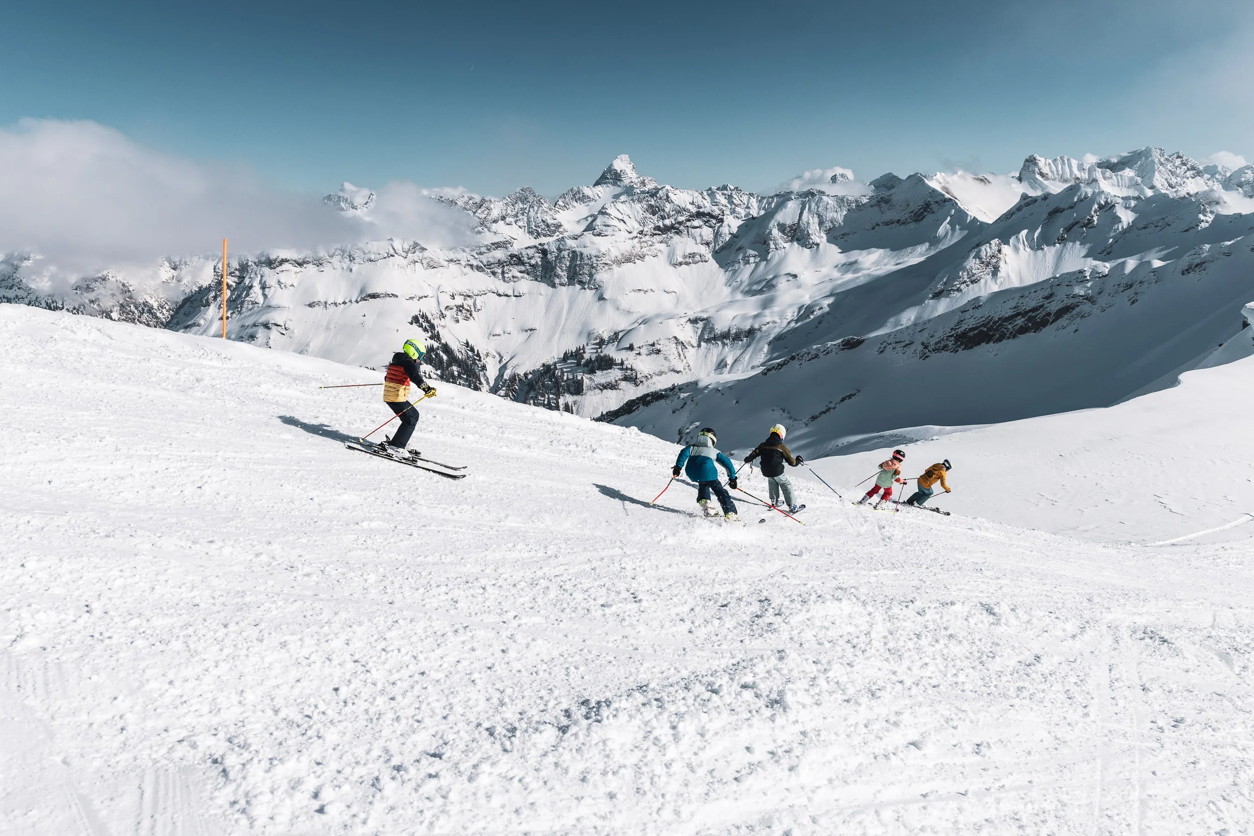 Kinderskigruppe fährt über eine breite Piste vor verschneitem Alpenpanorama