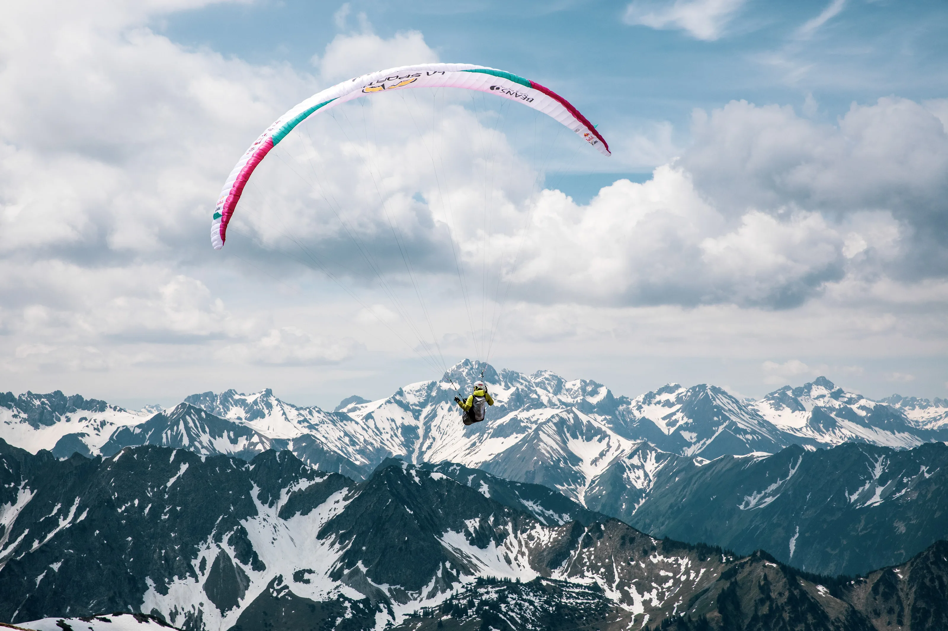 Michael Lacher fliegt unter dem großen pinkweißen Gleitschirm vor der verschneiten Alpenkette.