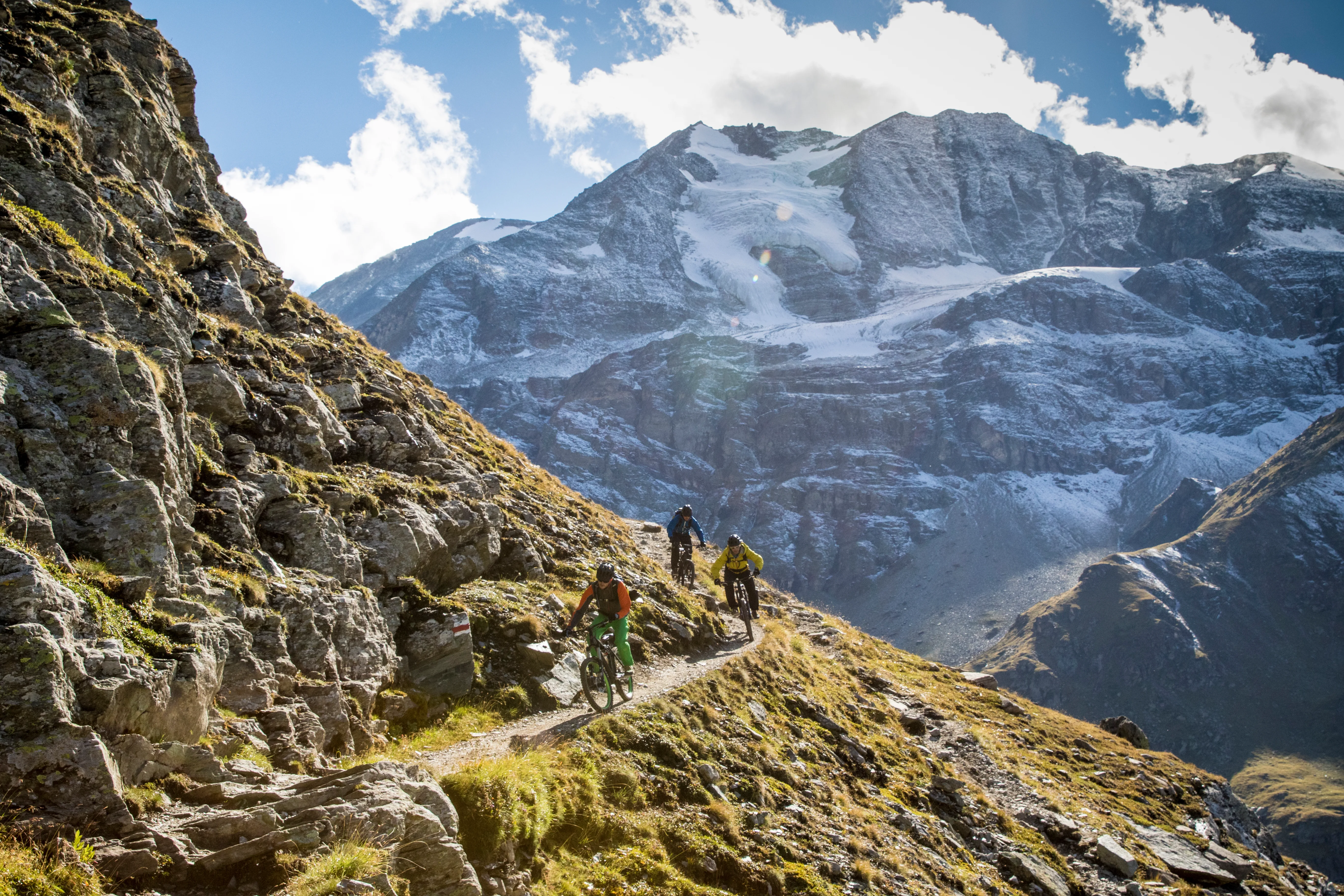 Robert Härle, Florian Bergmann und Florian Häusler fahren auf einem schmalen Bergtrail vor vergletscherten Gipfeln.