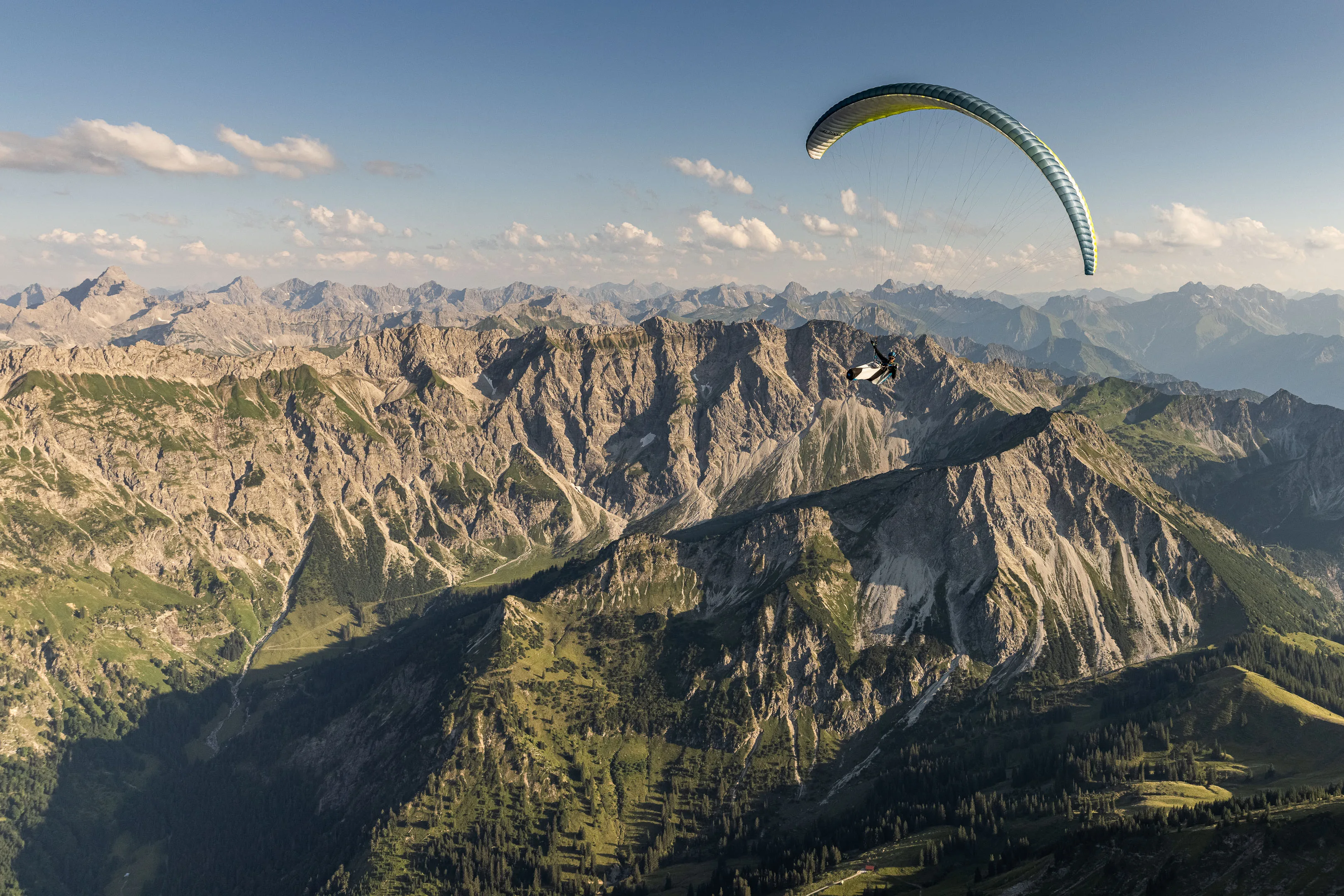 Ein Gleitschirmpilot fliegt vor steilen Flanken und grünem Gipfelgelände
