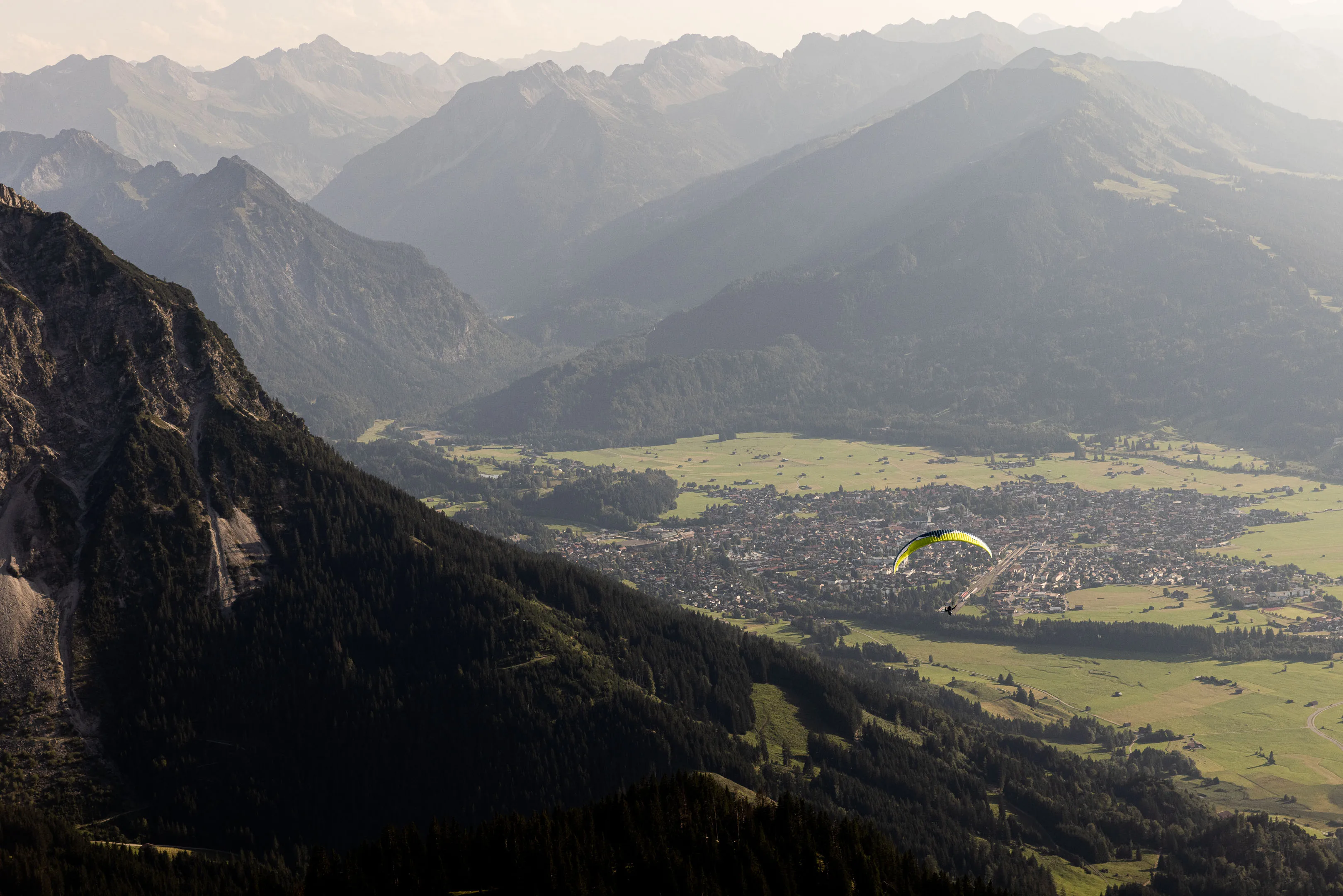 Ein Gleitschirm zieht über Oberstdorf durch den Schatten steiler Bergflanken