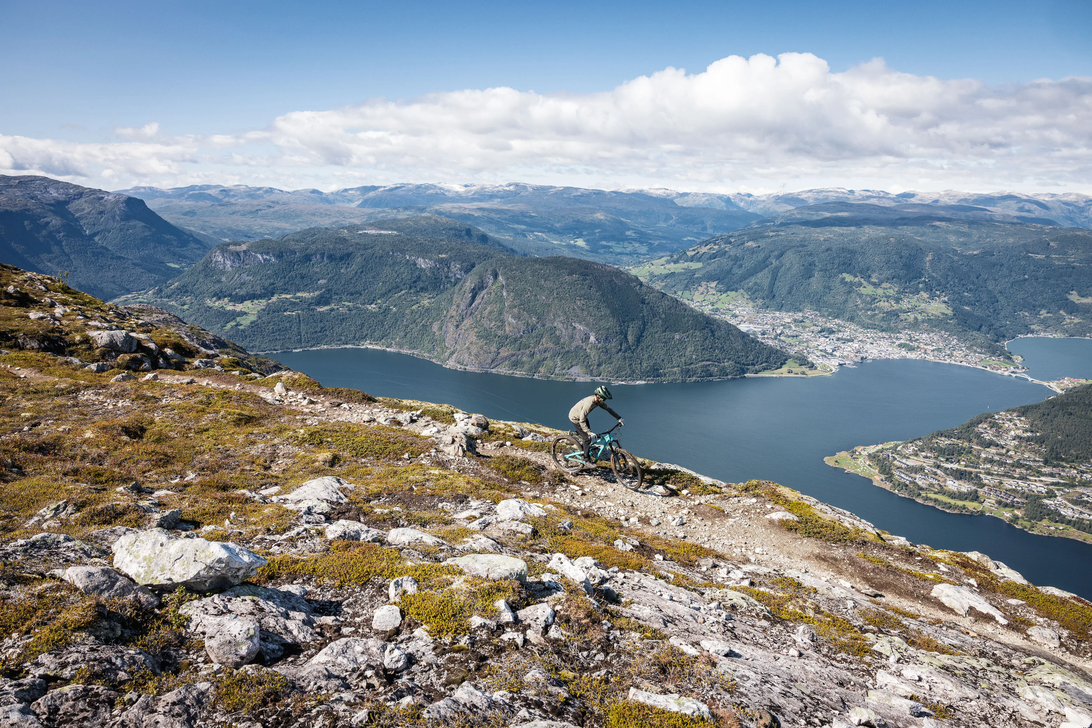 Mountainbiker fährt einen felsigen Hang mit Blick auf Fjord und Siedlung hinab
