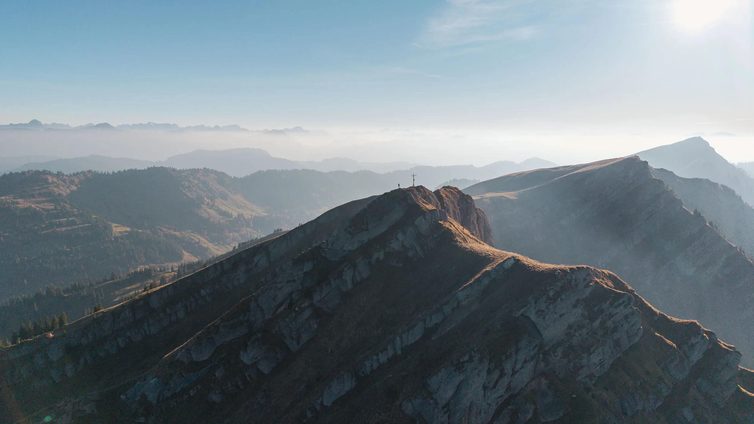 Ein felsiger Gipfelgrat mit Kreuz liegt vor einer weiten, dunstigen Berglandschaft