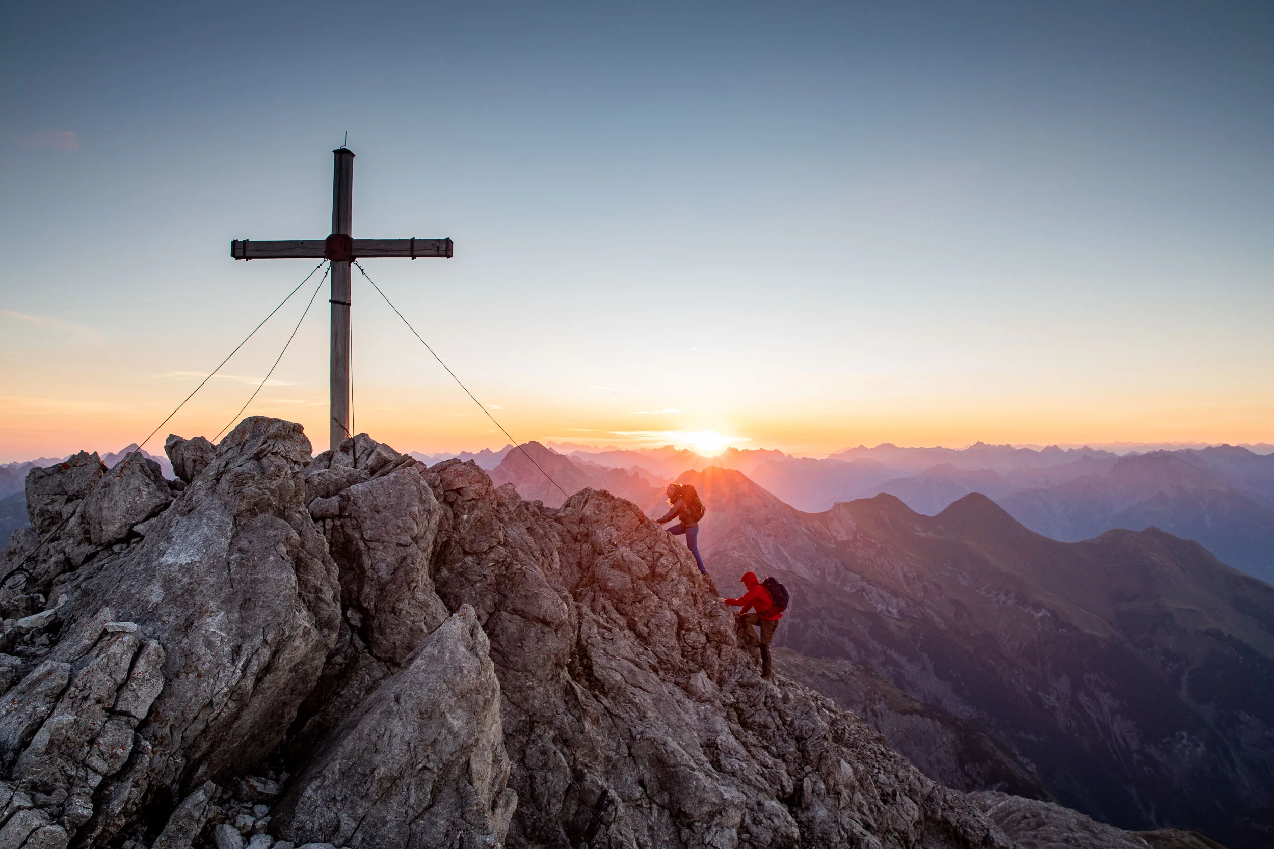 Ein Gipfelkreuz ragt über schroffe Felsen vor einer tief stehenden Sonne