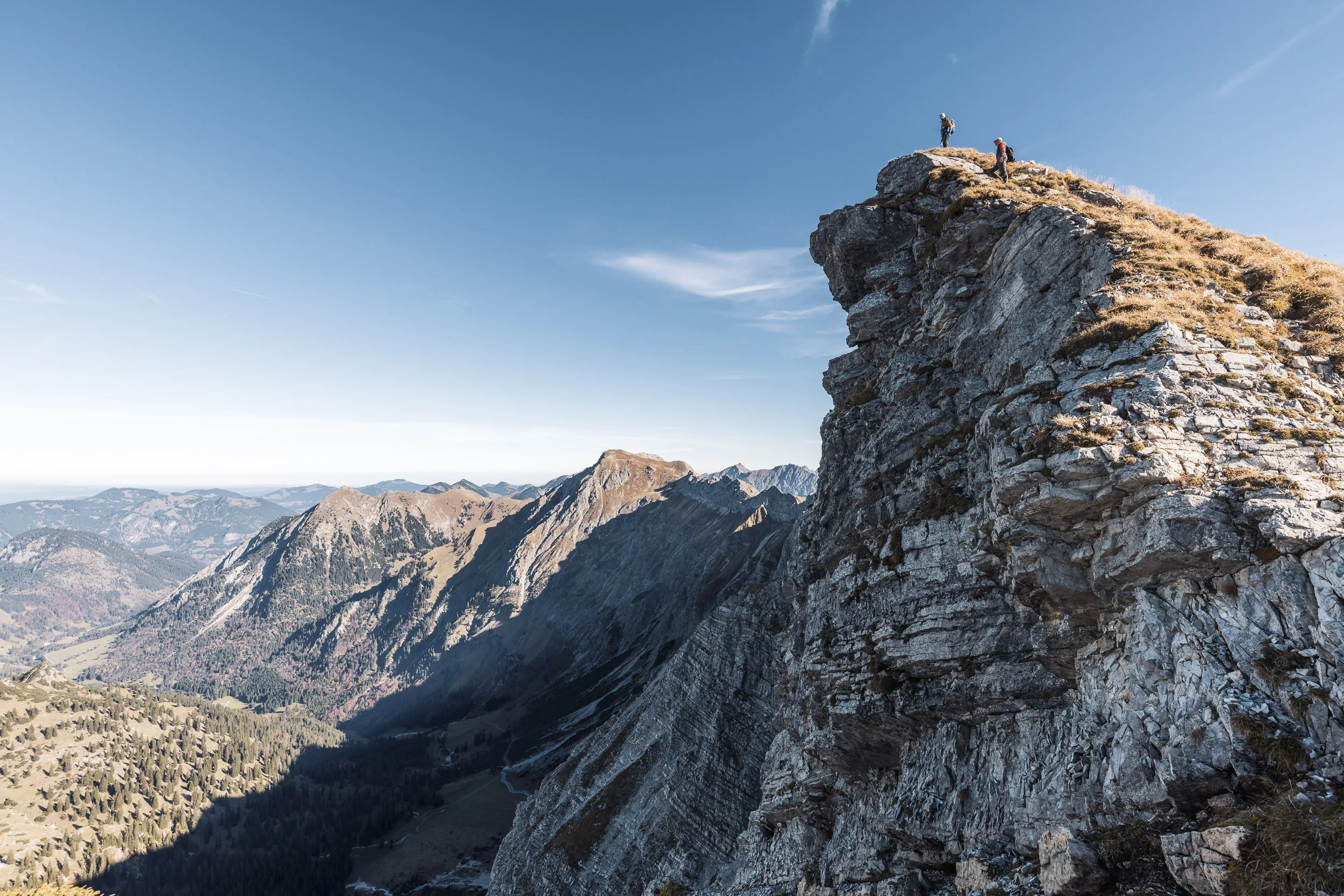 Zwei Bergsteiger stehen auf einem hohen Felsgrat über dem herbstlichen Tal.