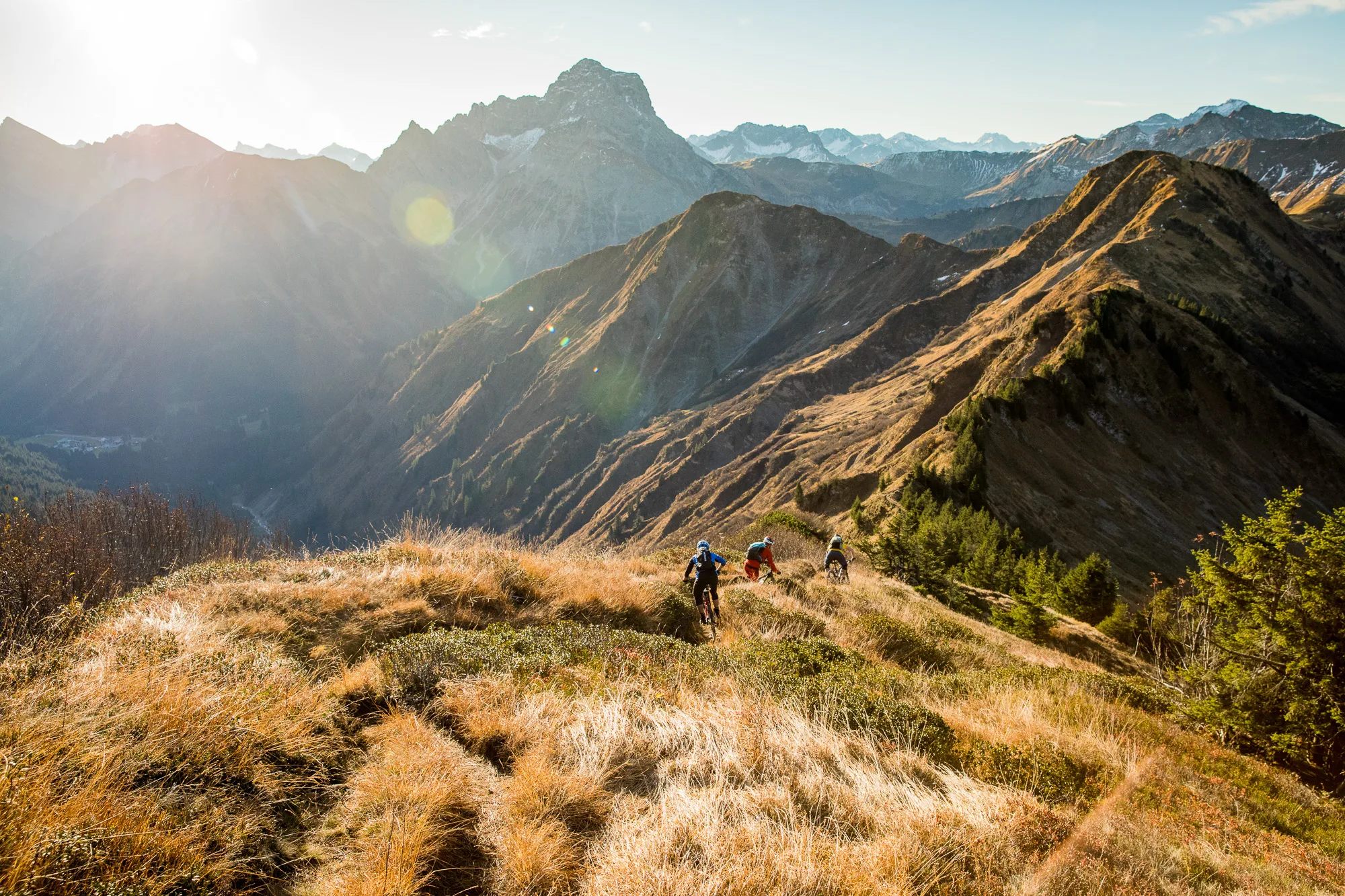 Mountainbiker queren eine helle Bergwiese mit dem Kleinwalsertal im Gegenlicht