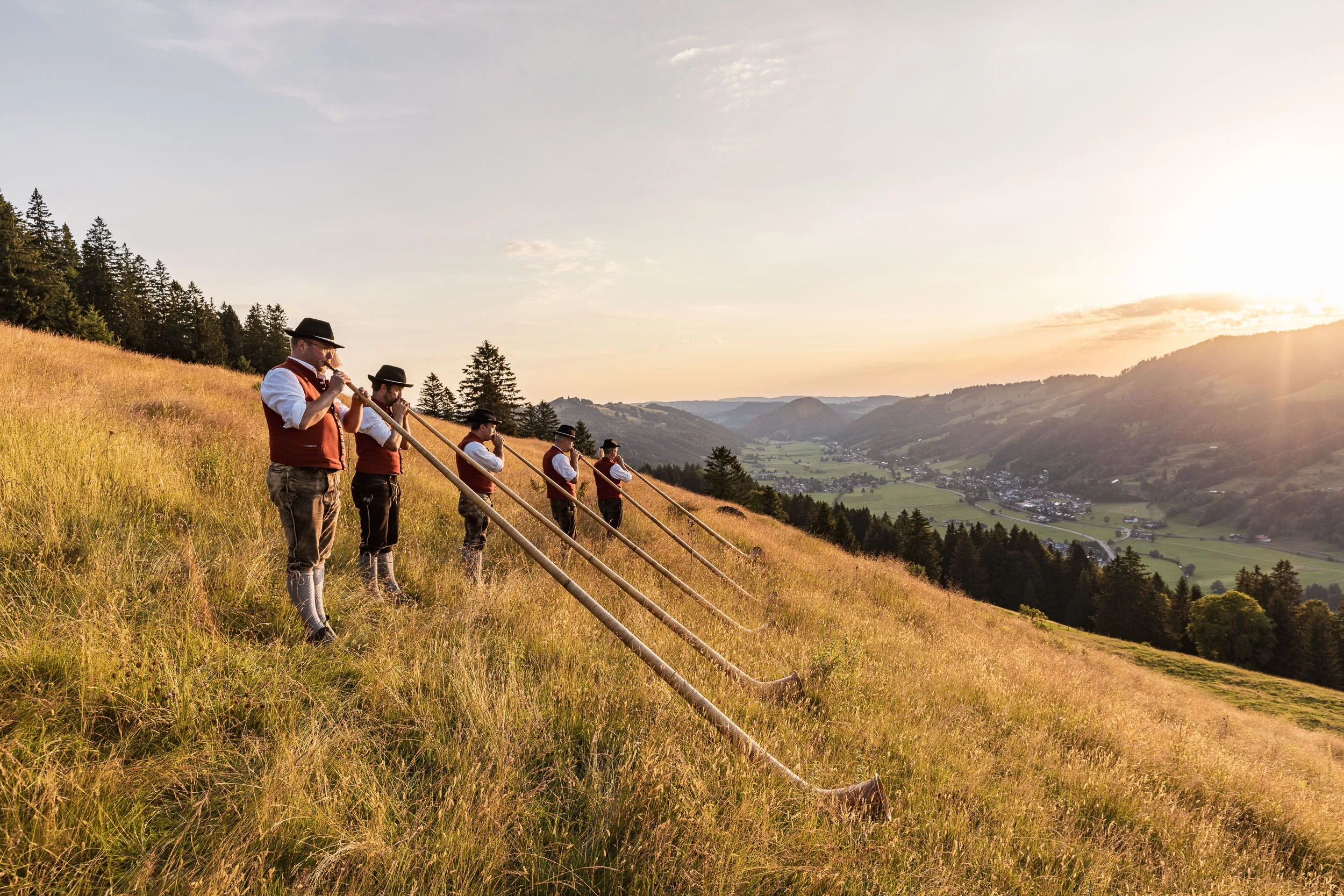 Alphornbläser auf einer goldenen Bergwiese im Allgäu kurz vor Sonnenuntergang.