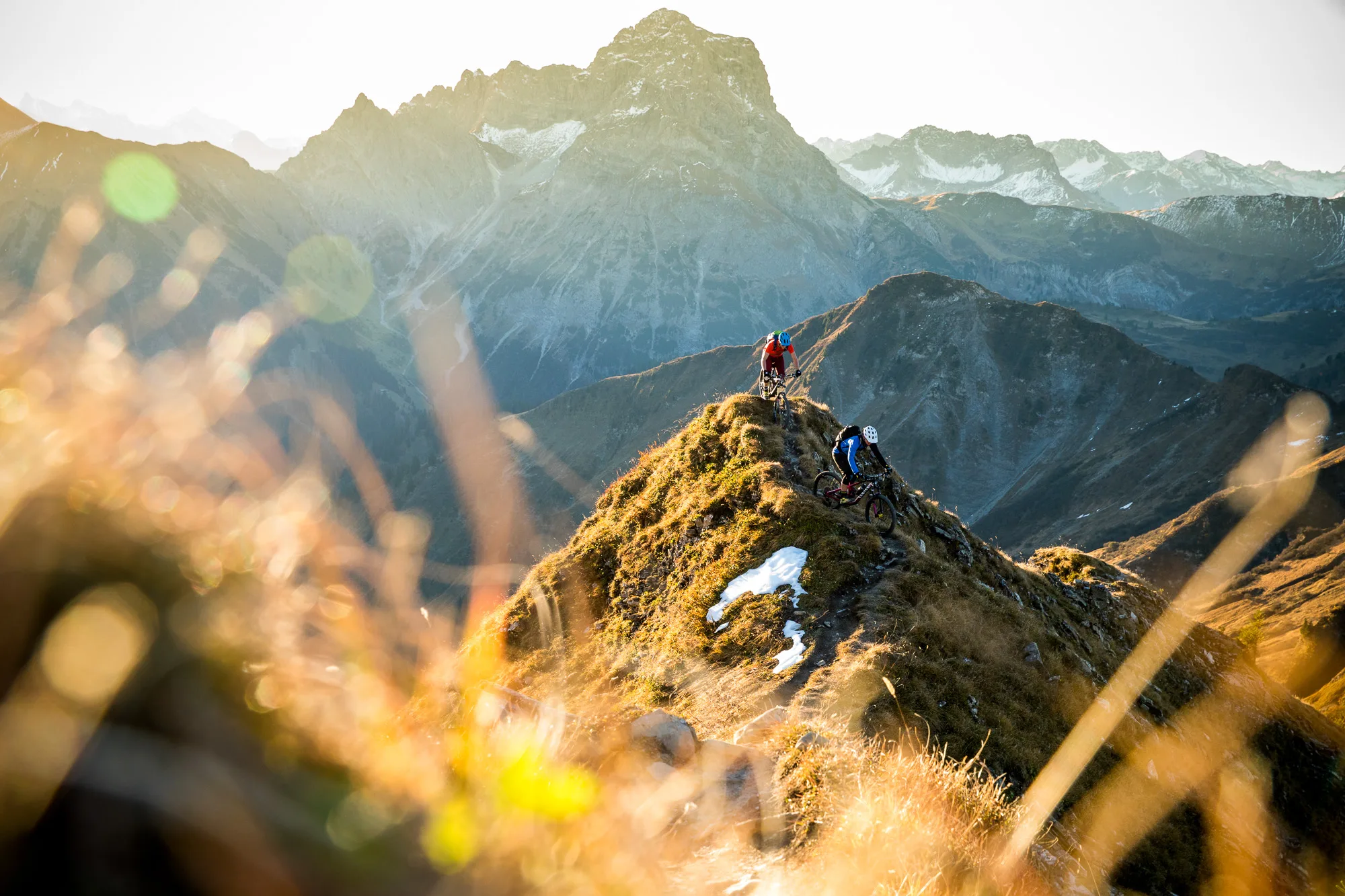 Zwei Mountainbiker bewegen sich über einen schmalen Grat vor einem weiten Alpenpanorama