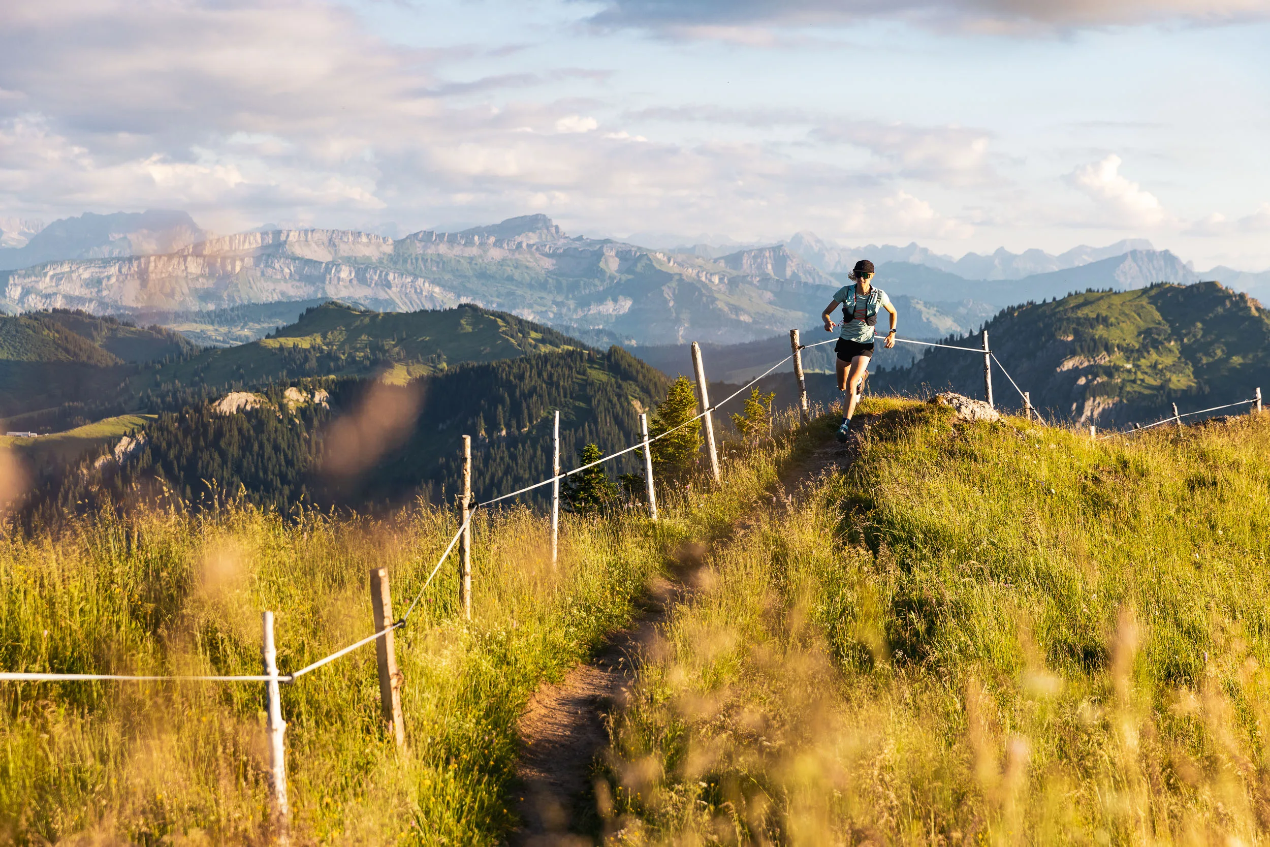 Eine Trailrunnerin läuft auf einem schmalen Gratweg an Zaunpfählen vorbei, dahinter liegt ein weites Bergpanorama.