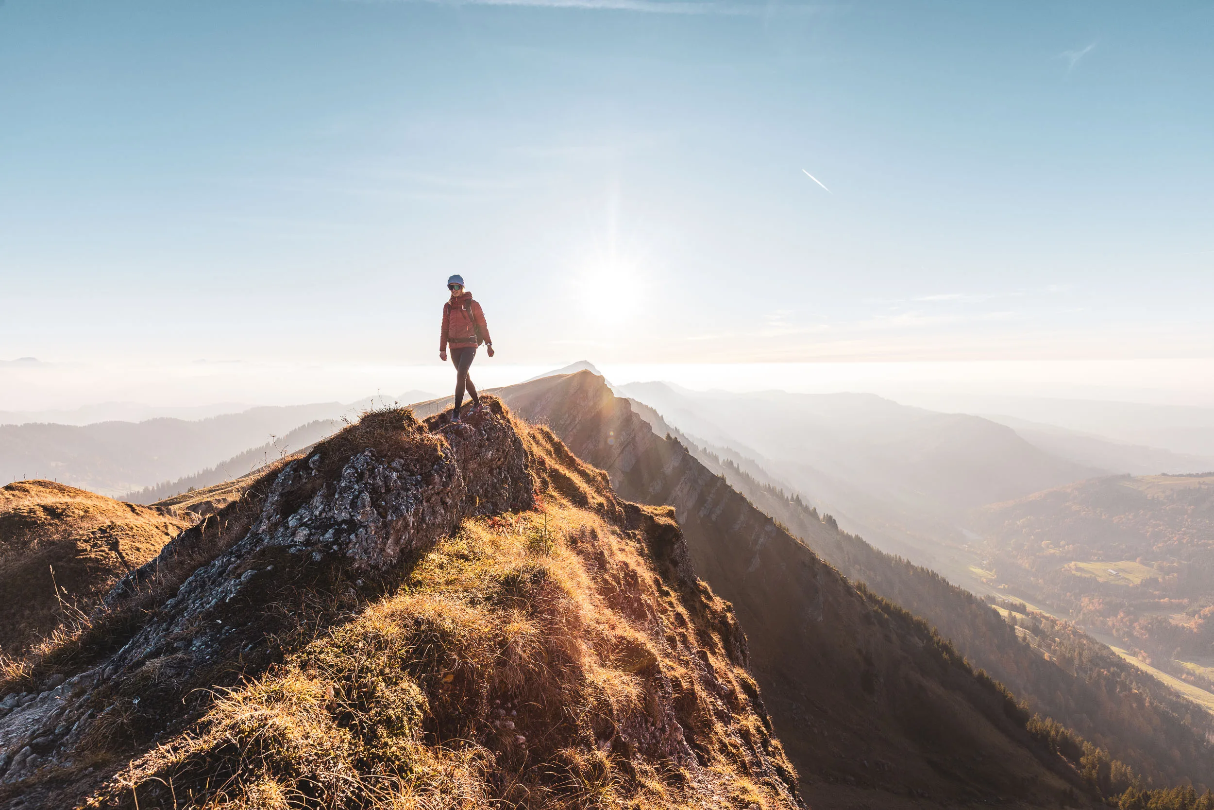 Wanderin steht klein auf einem Grat vor einem breiten, dunstigen Bergpanorama