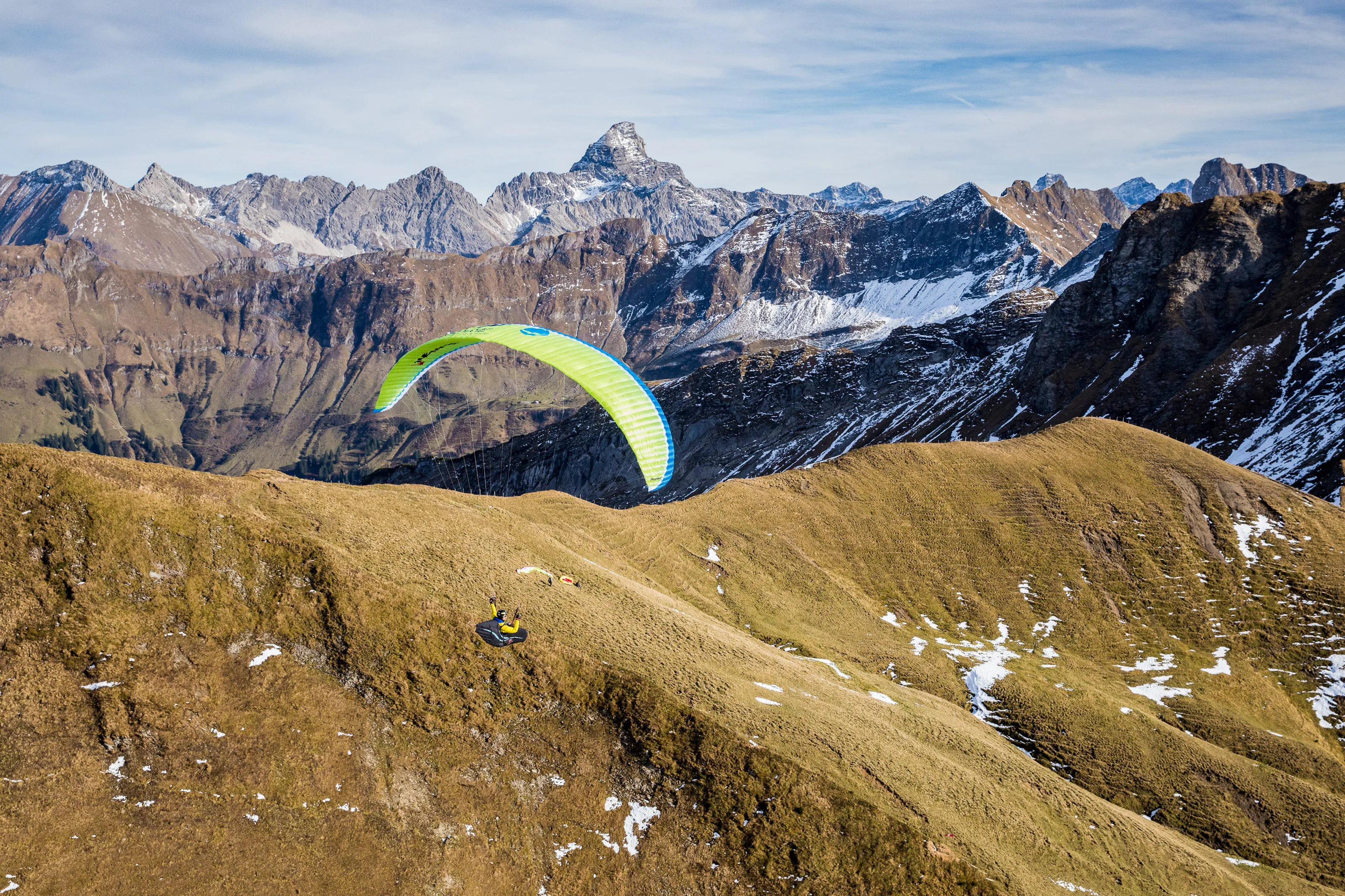 Michael Gebert fliegt mit grünem Gleitschirm über eine goldene Bergflanke vor schneebedeckter Alpenkulisse