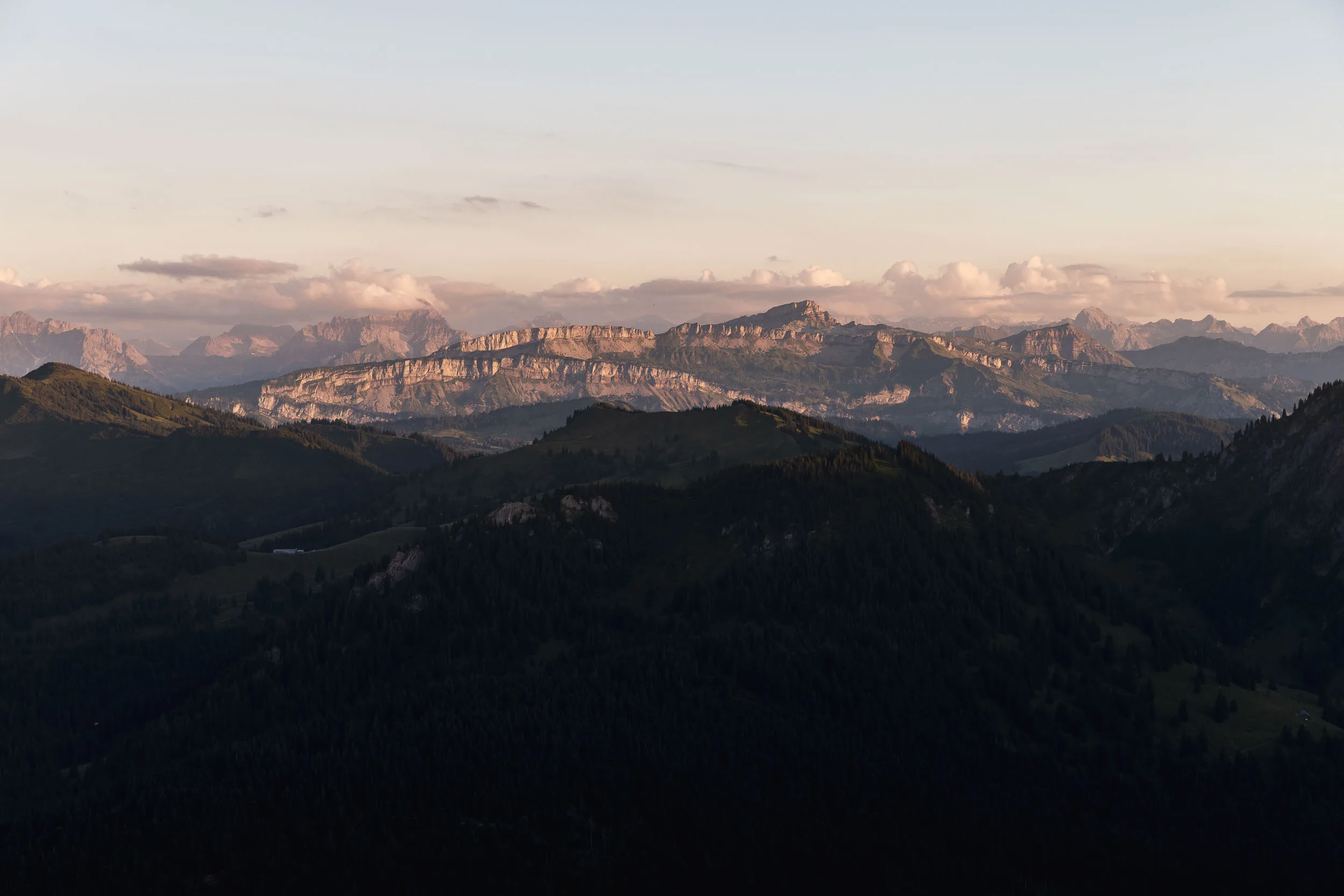 Ferne Felswände leuchten im Abendlicht über dunklen, bewaldeten Bergrücken.